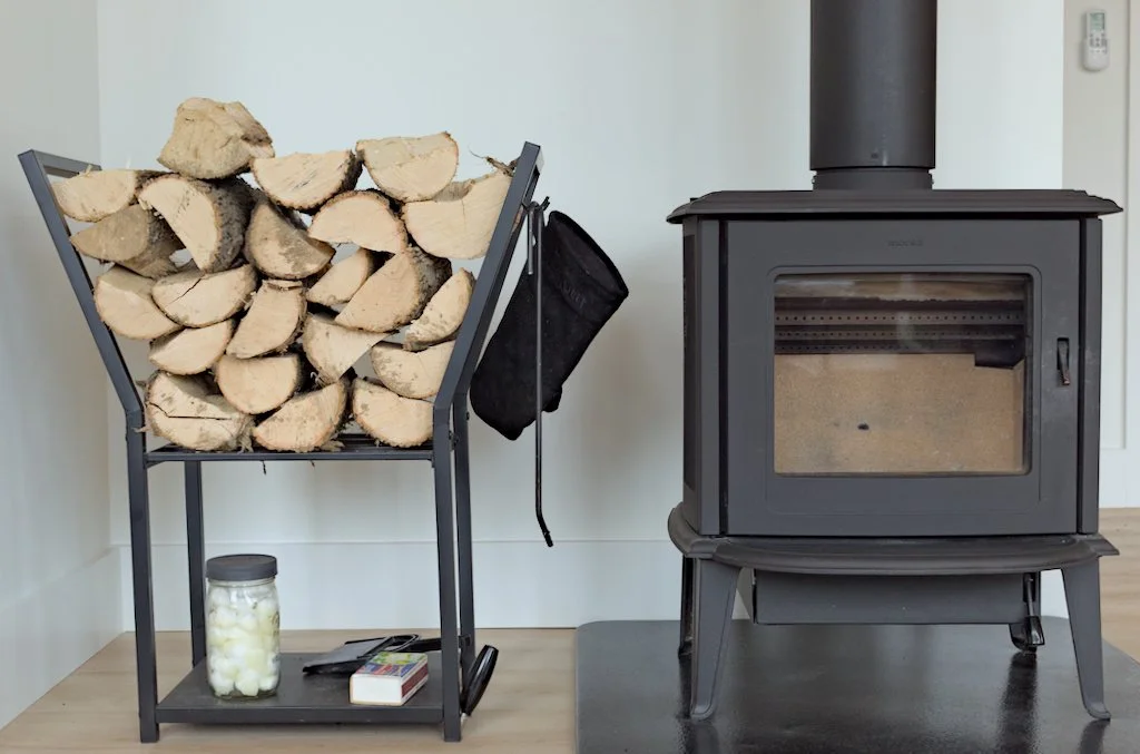 Wood stove with metal log rack filled with firewood and kindling container beside it.