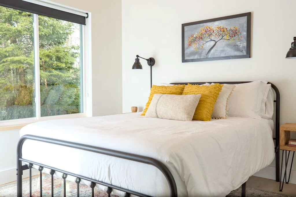 Modern bedroom with a black metal bed frame, white bedding, and yellow accent pillows, near a large window. Wall art depicting a colorful tree is hung above the bed. Two black wall-mounted lamps flank the bed.