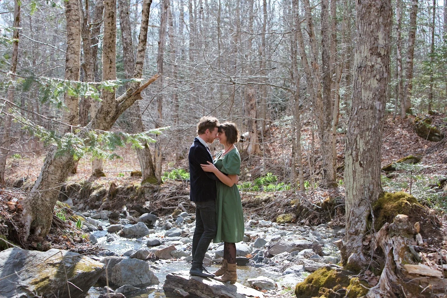 A couple standing on rocks in a forest, touching foreheads and embracing near a small stream surrounded by trees.