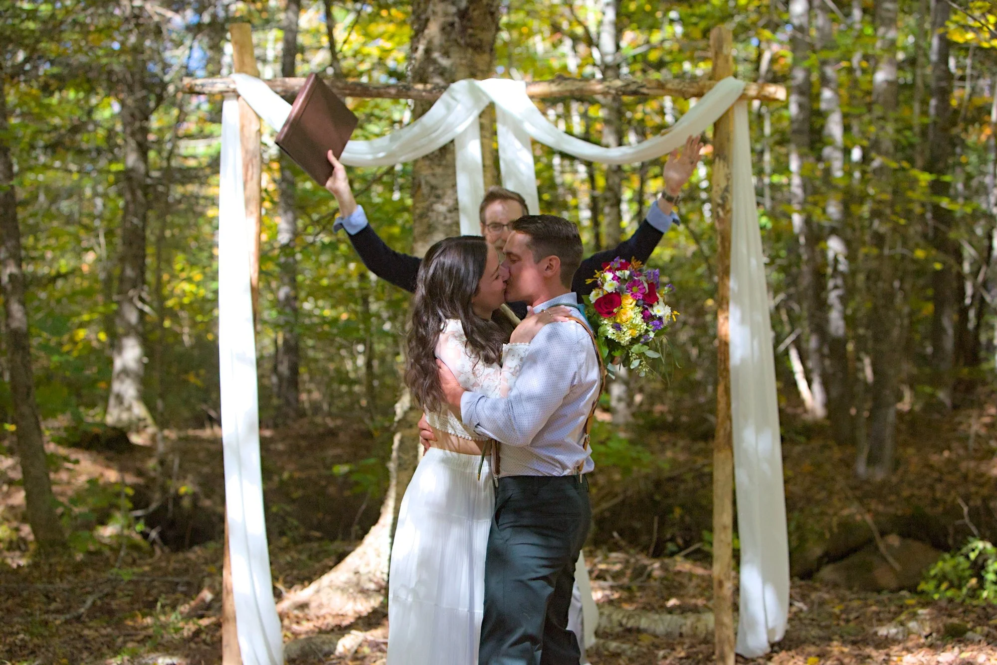 A couple kiss during a wedding ceremony in the woods, with an officiant standing behind them. The couple is holding a bouquet of flowers, and there is an arch decorated with white fabric and wood.