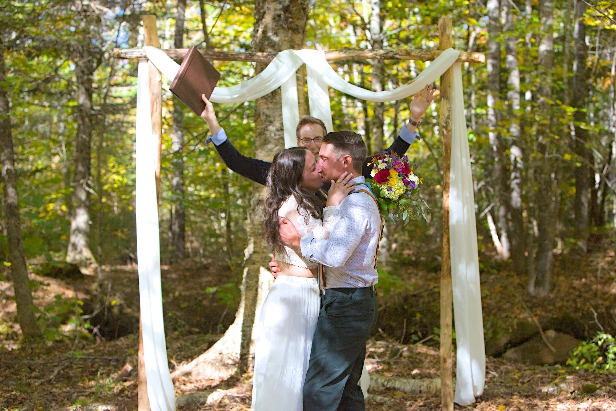 A couple kissing during their wedding ceremony in a forest, with an officiant standing behind them, holding a book and raising his hand. The ceremony is decorated with white fabric and a bouquet of colorful flowers.