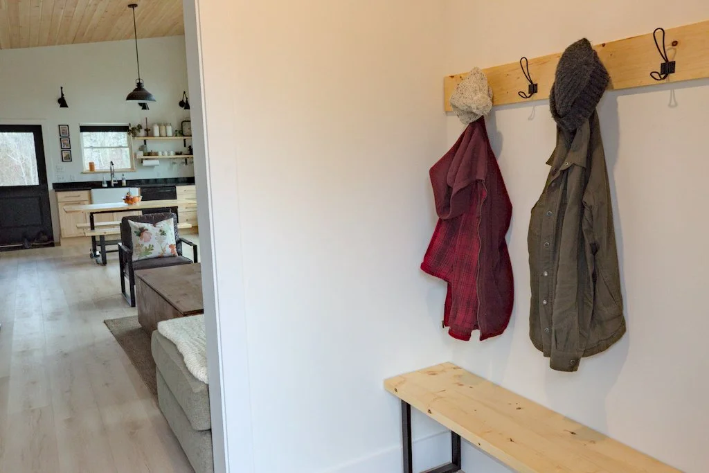 Interior of a home with a mudroom area featuring coats and hats on hooks, a bench, and a view of a kitchen and living room with light wood floors, pendant lights, and shelves.