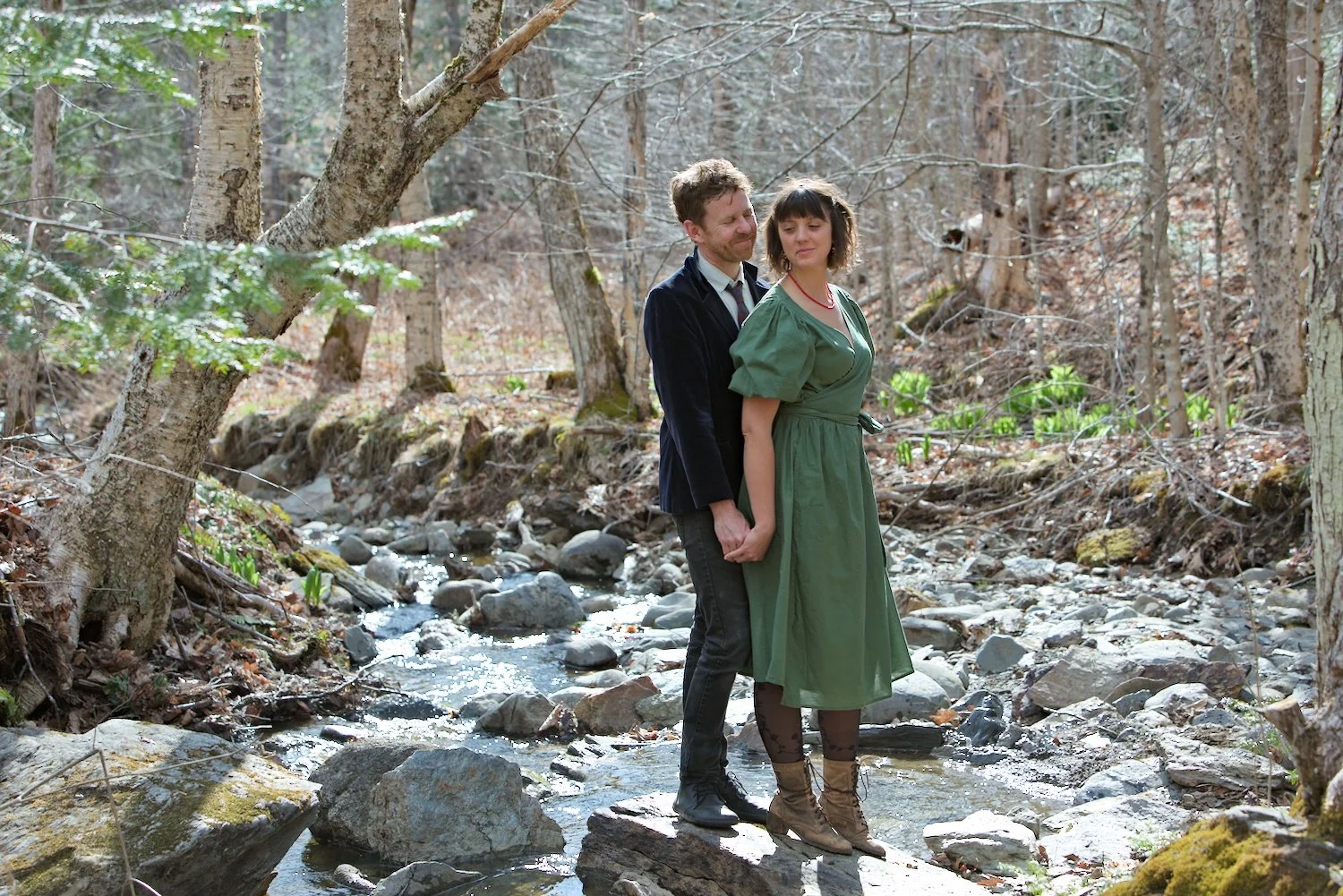 A man and woman holding hands standing on rocks in a brook in a forest