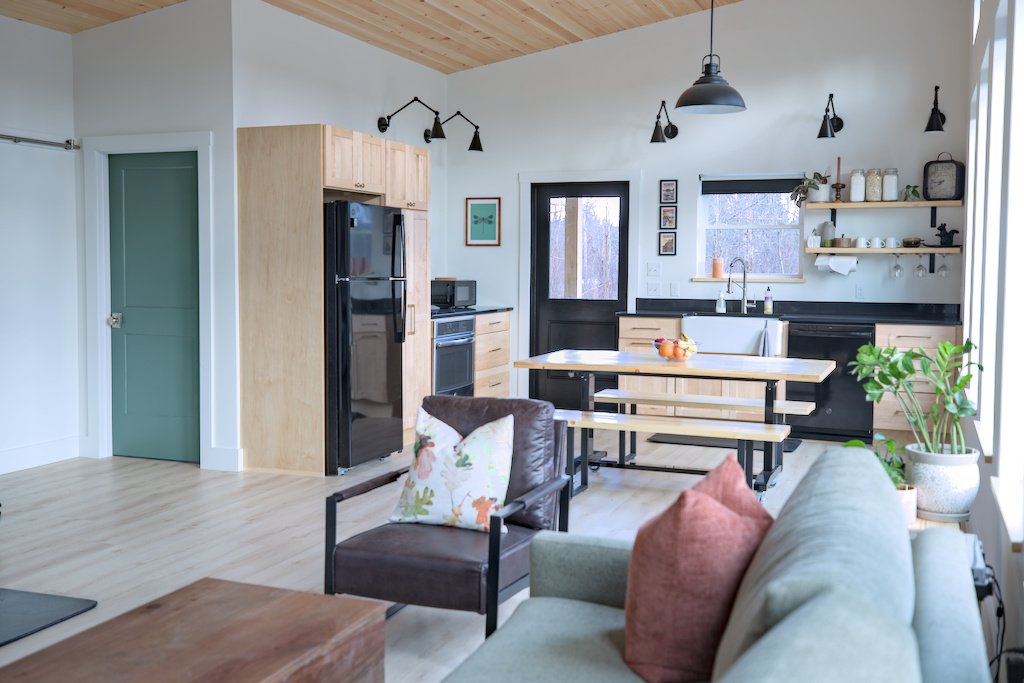 Modern open-concept kitchen and living room with light wood finishes, green door, black appliances, and indoor plants. Features a wooden table with benches, a sofa, and decorative lighting.