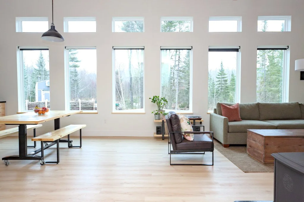 Modern living room with large windows, green sofa, wooden coffee table, armchair, indoor plant, and a dining table with benches. Light wood flooring and pendant light.