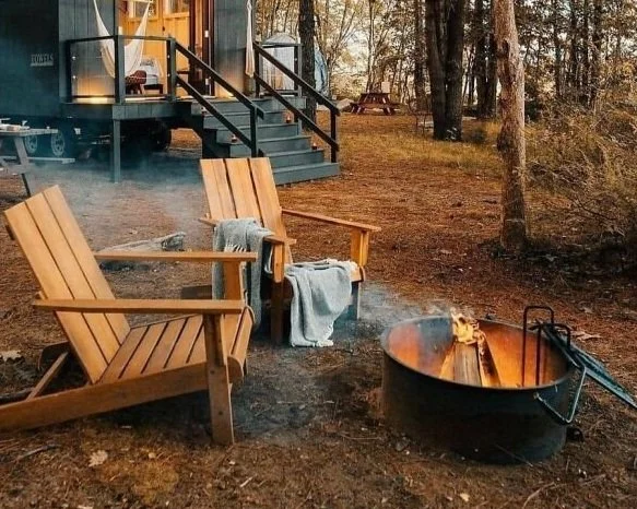Outdoor wooden chairs around a fire pit in a forested area with a cabin in the background. Towels are draped over the chairs, and a gentle fire is burning, emitting smoke.