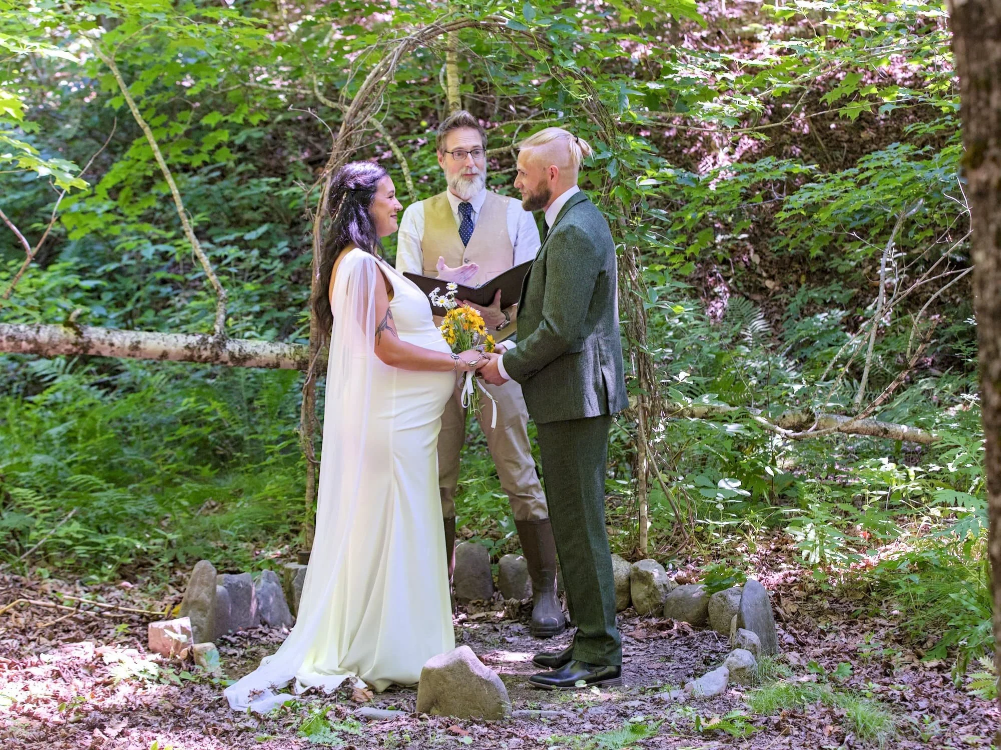A couple is getting married outdoors in a wooded area, holding hands and facing each other, with a woman in a white dress and a man in a suit. An officiant stands behind them, holding a book, under a rustic arch made of branches. The setting is lush and green, with sunlight filtering through the trees.