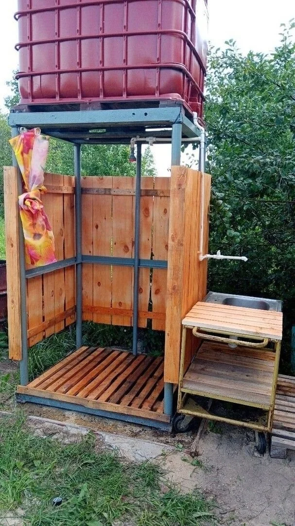 Outdoor shower setup with wooden enclosure, metal frame, and large water tank on top, adjacent to a small sink with a wooden counter.