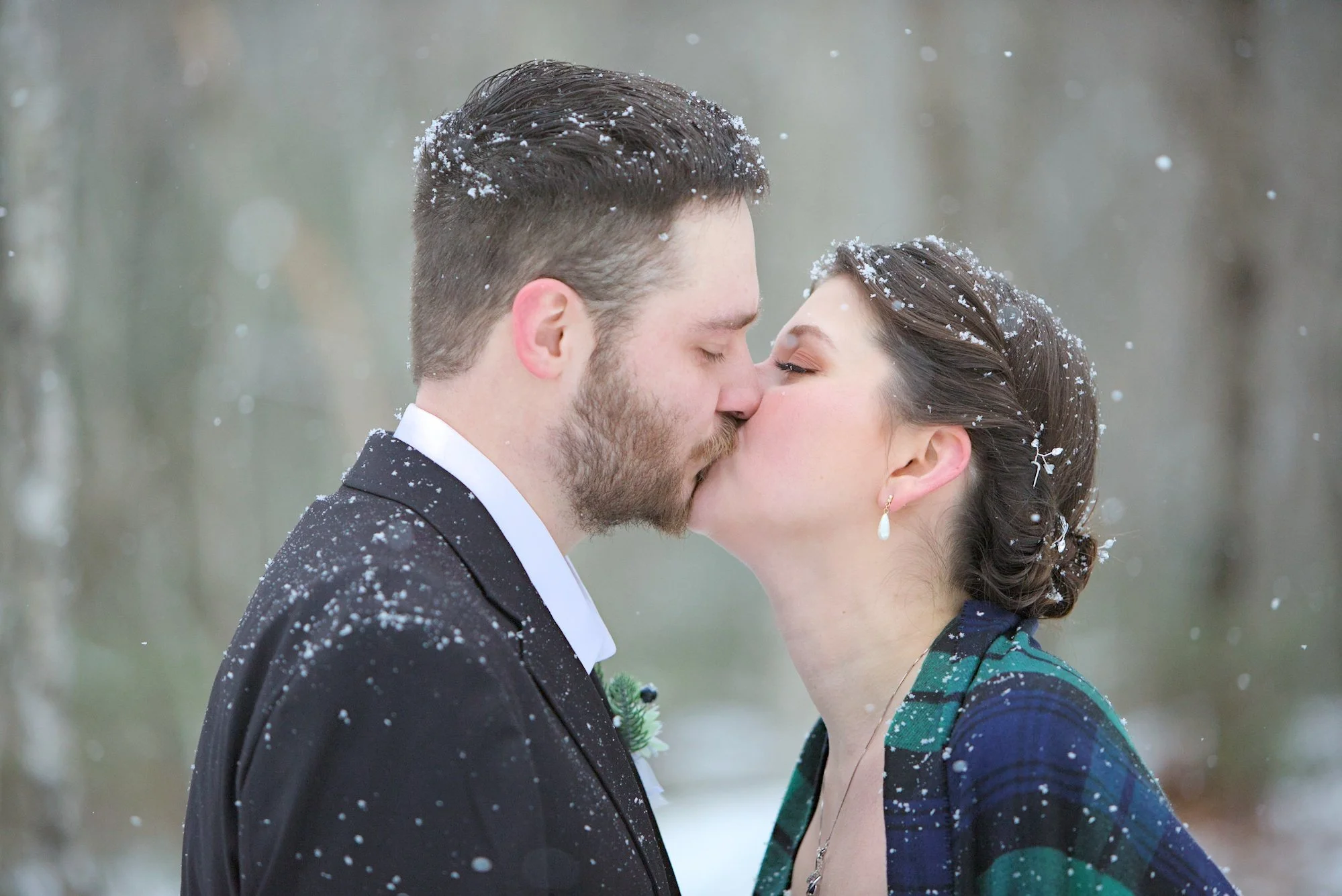 A man and woman share a kiss in a snowy outdoor setting, both with their eyes closed. Snowflakes are on their hair and clothing.