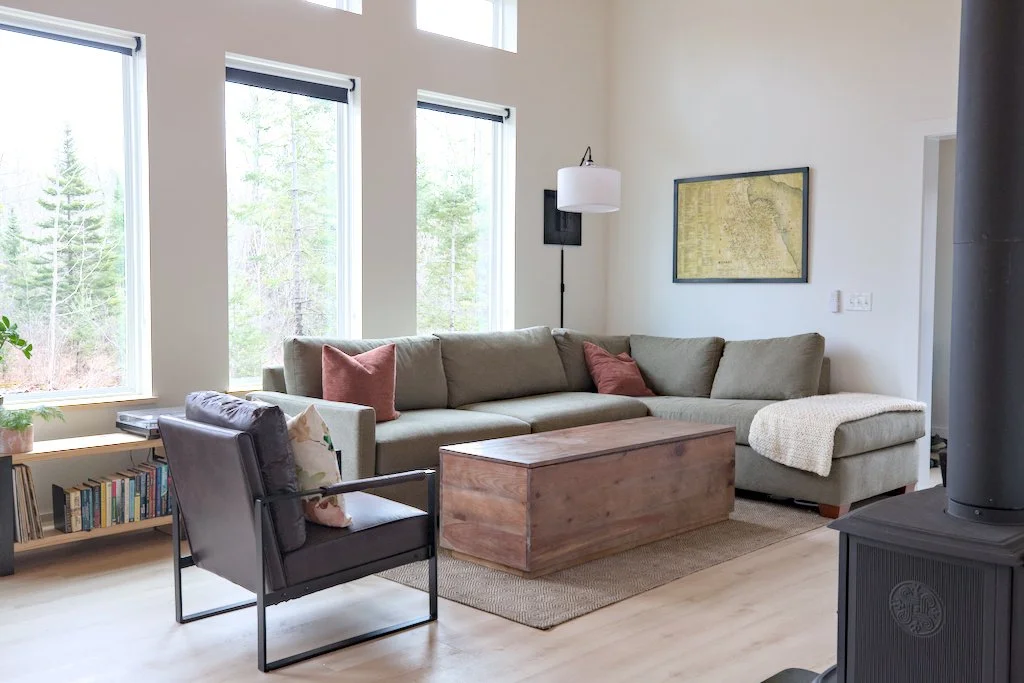 Cozy living room with large windows, corner sofa, armchair, wooden coffee table, wall art, and bookshelf.