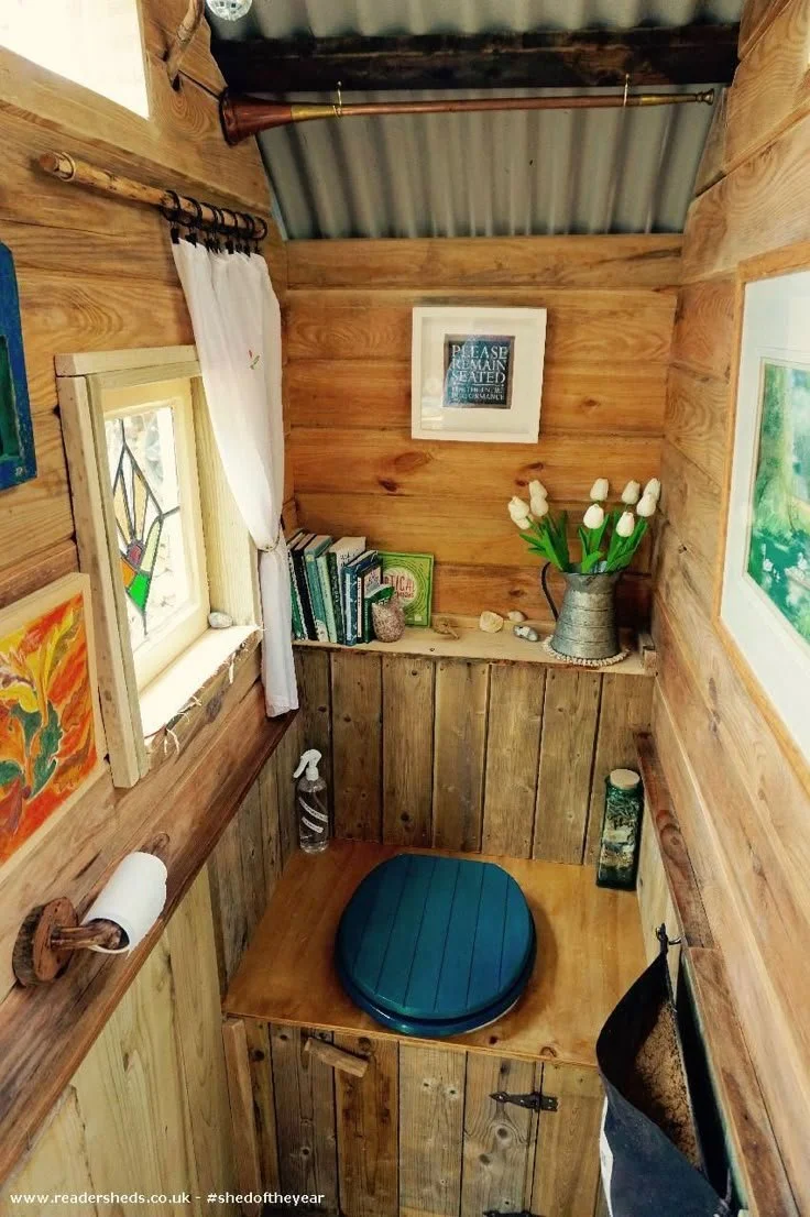 Compact wooden bathroom with a blue toilet seat, books, decorative items, tulips in a metal pitcher, and framed art on the walls.