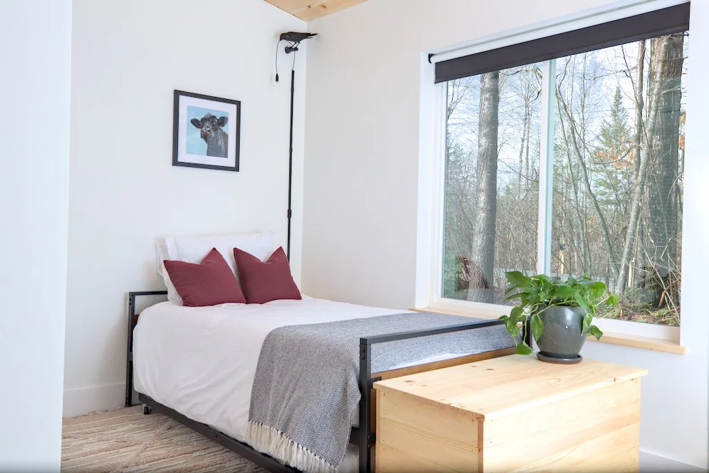 Minimalist bedroom with a single bed, white bedding, and burgundy pillows. A gray blanket is at the foot of the bed. A small wooden chest with a potted plant sits near a large window with a view of trees. A picture hangs on the wall above the bed.