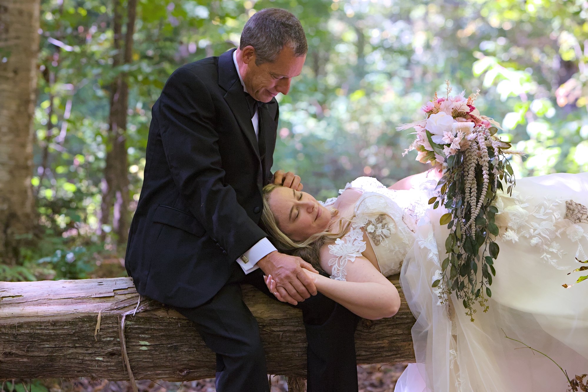 A man in a black suit is holding and leaning over a woman lying on a log in a forest during daylight. The woman is wearing a white lace wedding dress and is resting her head on the log. She has a relaxed expression, and there is a large bouquet of pink and white flowers with greenery on her chest. The forest background is blurred with green foliage and sunlight filtering through the trees.