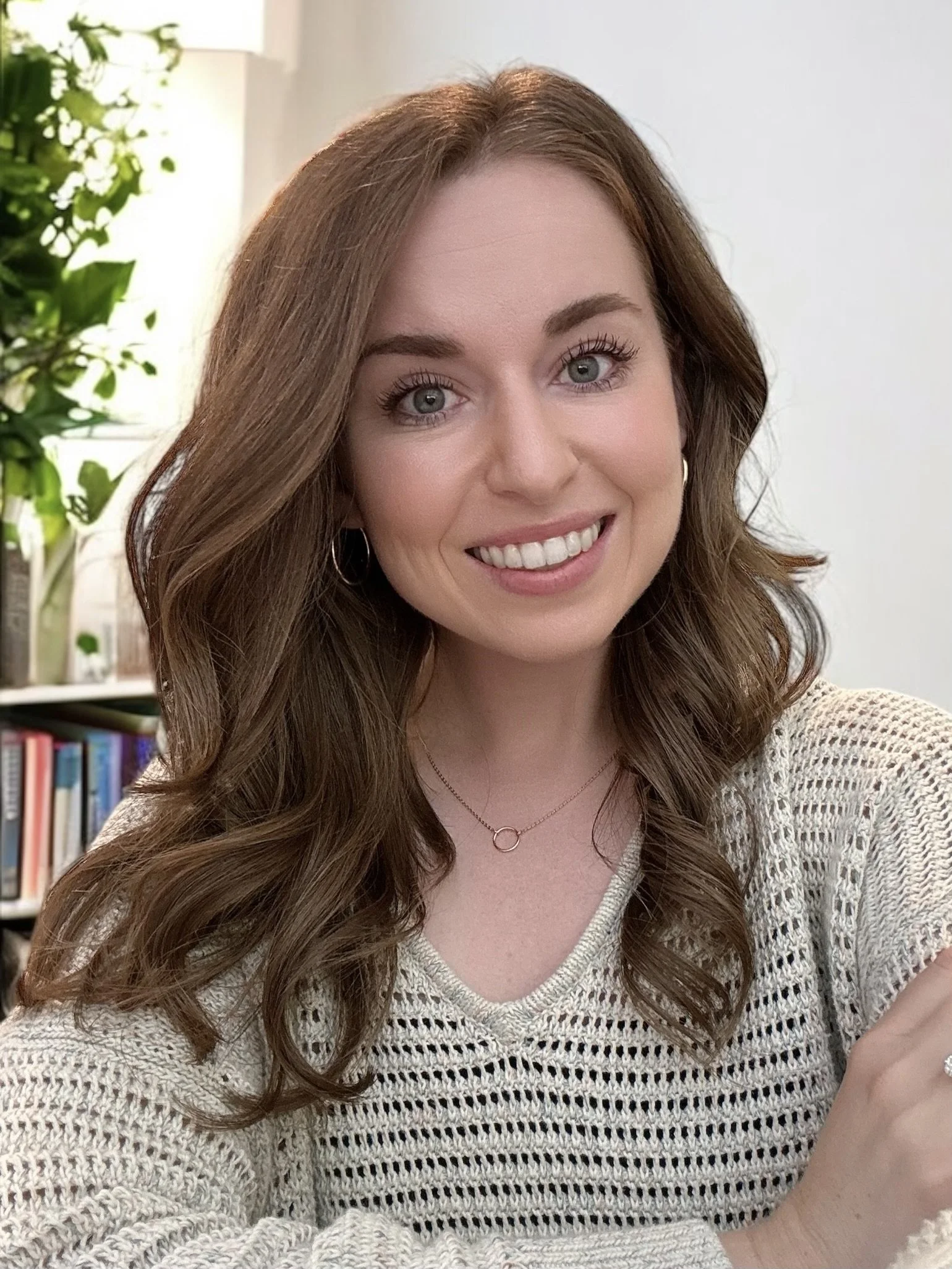 Smiling woman with long brown hair wearing a beige knitted sweater, sitting indoors next to a bookshelf and green plants.