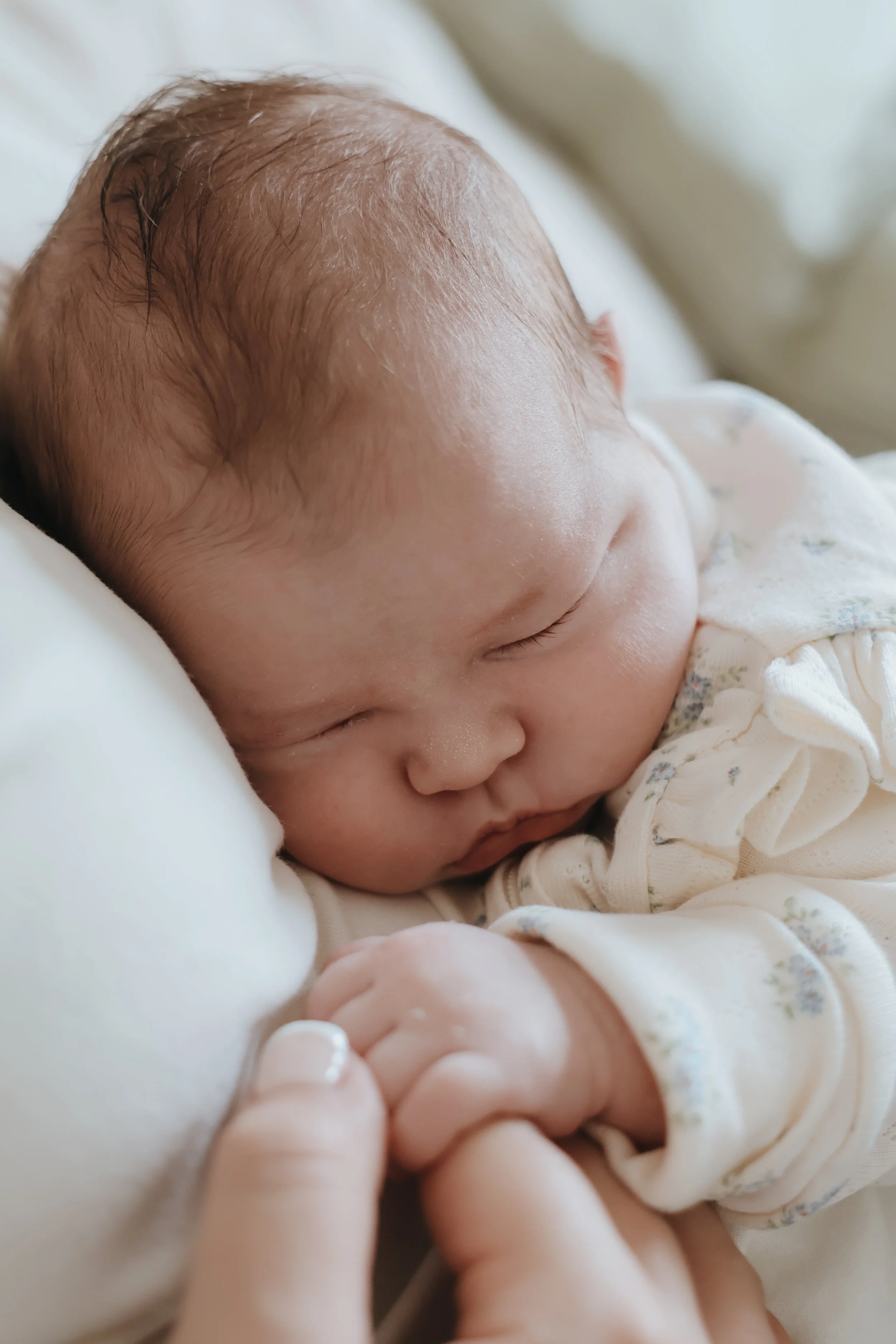 Close-up of a sleeping newborn baby with light-colored hair, lying on a white pillow, wearing a beige outfit with small floral patterns, gently holding a parent's finger.