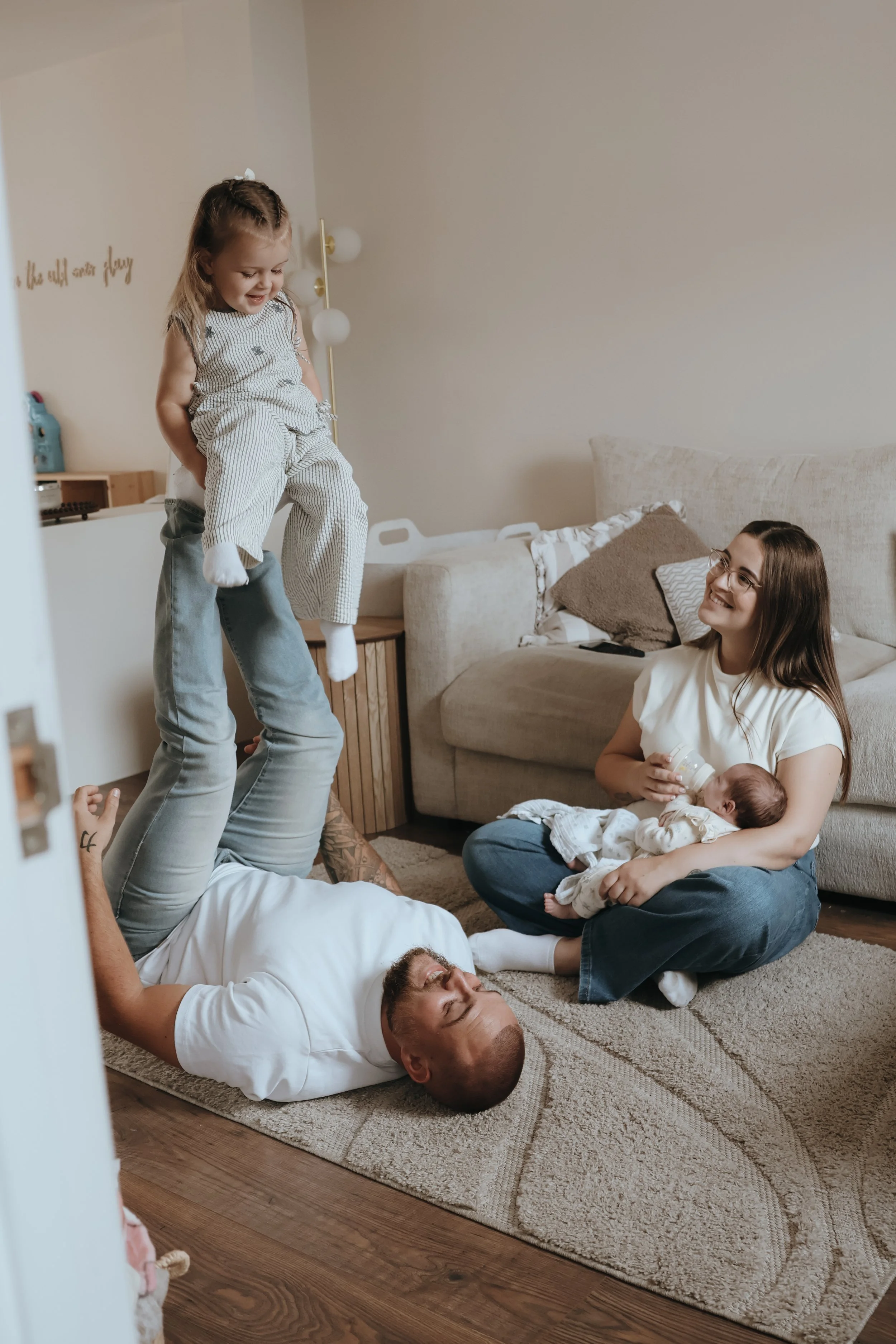 A group of family members, including children and adults, are playing and relaxing on the floor of a living room. One child is balancing on an adult's legs, while a woman sits on the floor holding a baby. The scene appears joyful and playful.