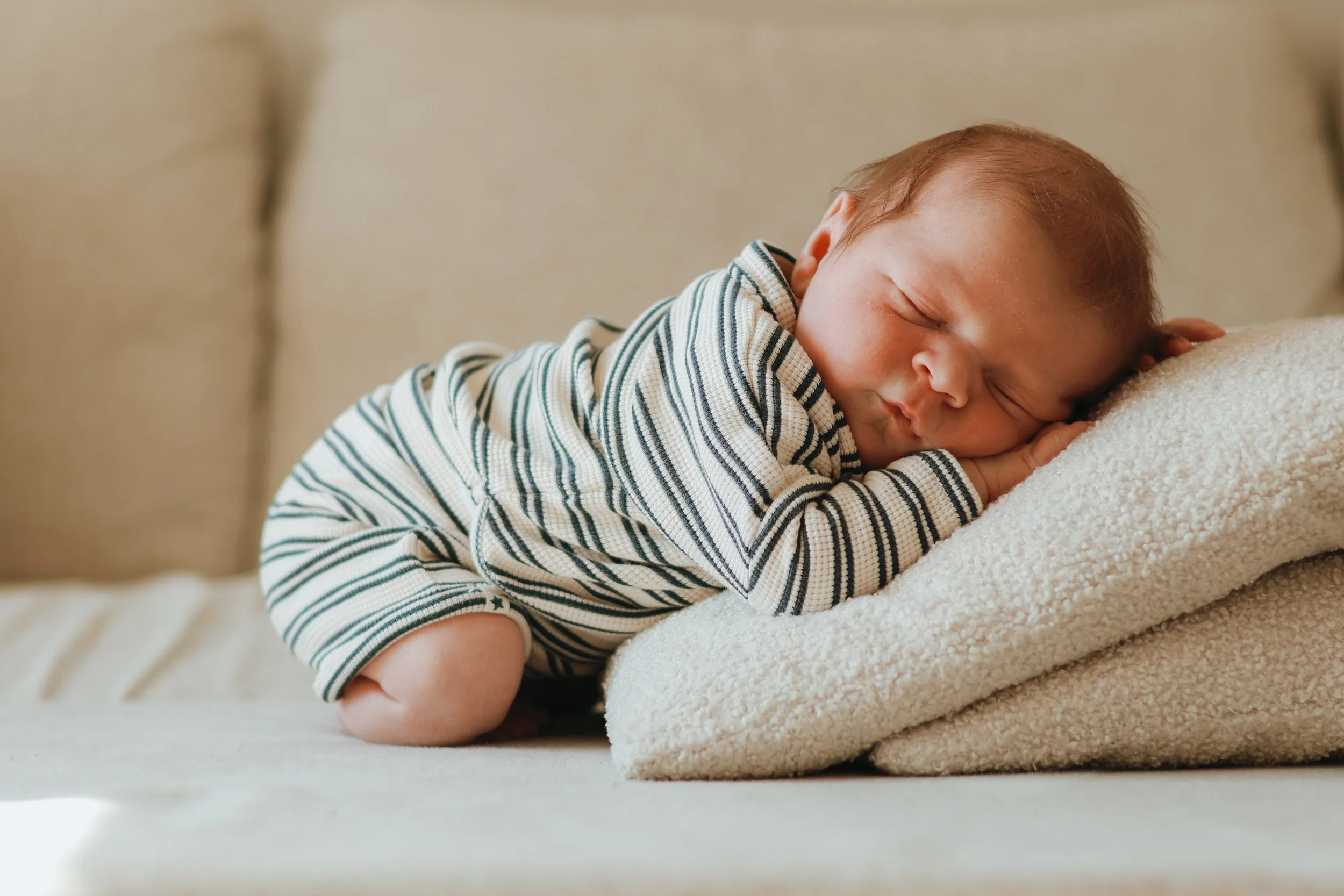 A young child with red hair sleeping peacefully on a beige couch, resting their head on a soft, cream-colored pillow while wearing a striped long-sleeve shirt.