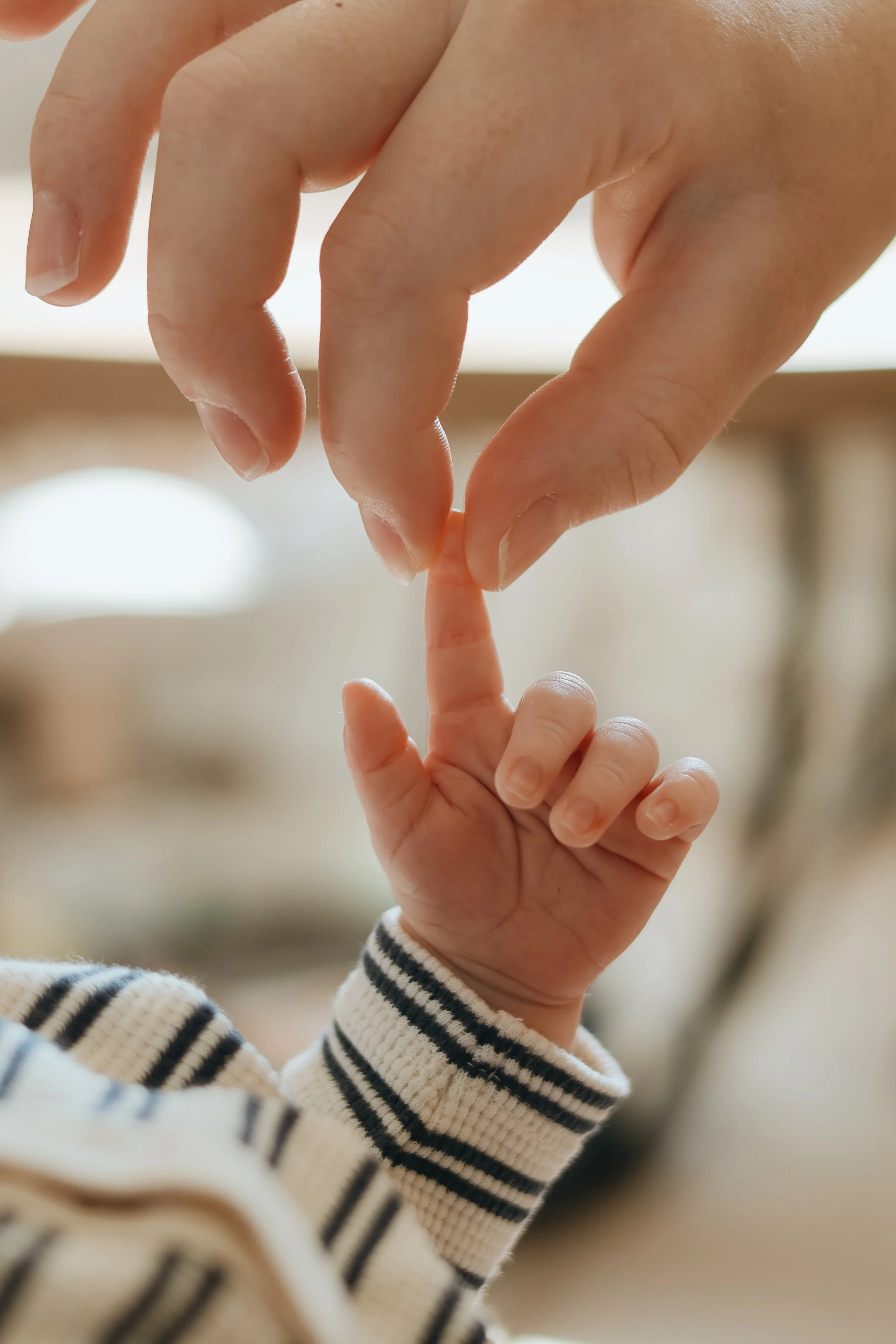 An adult hand gently holding a tiny baby finger, with the baby hand and finger in foreground and adult hand reaching down from above, in a close-up shot.