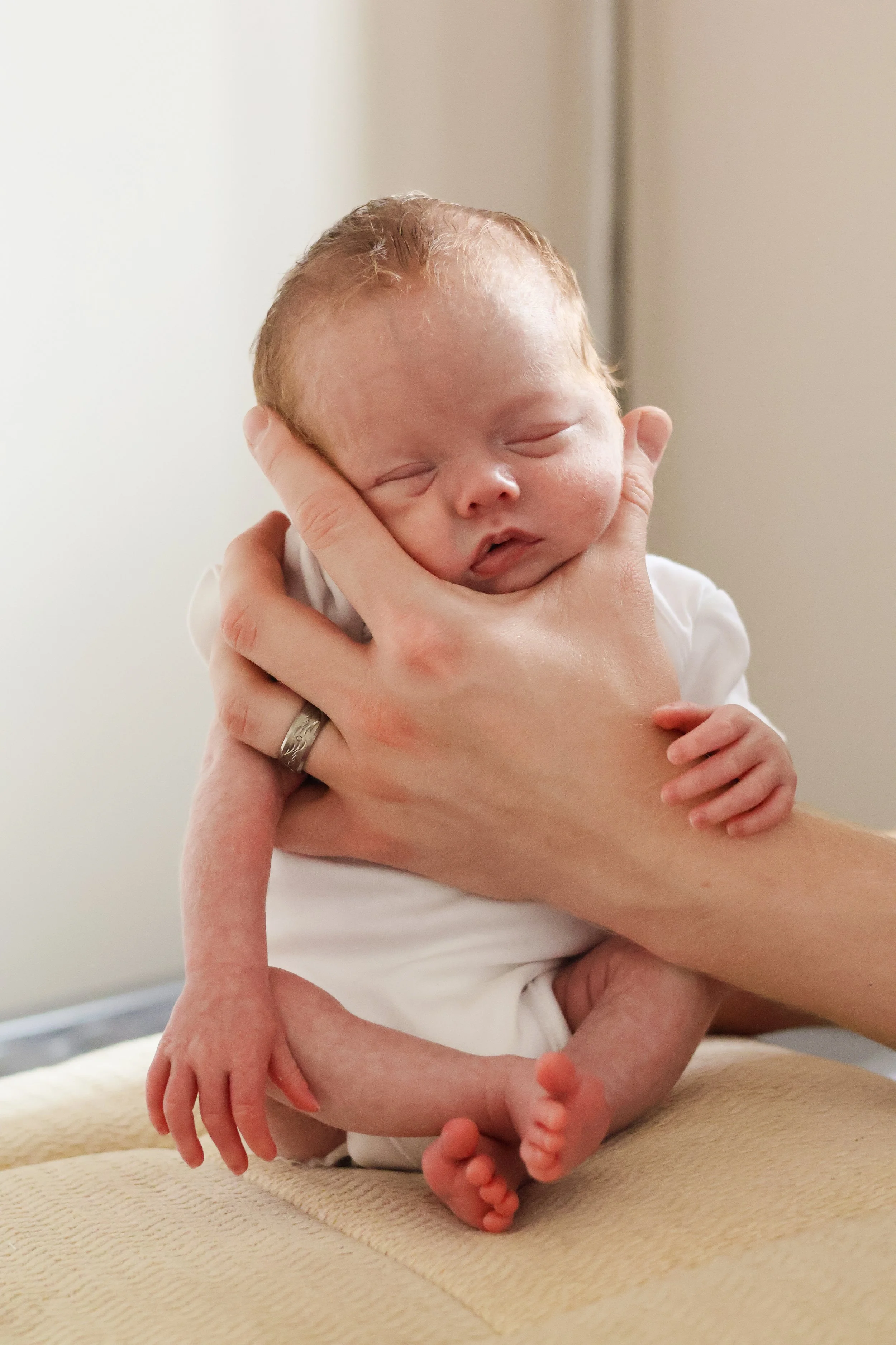 A person is holding a sleeping newborn baby with red hair, supporting the baby's head and body gently with one hand, while the baby's legs are crossed and feet are visible. The scene appears to be indoors.