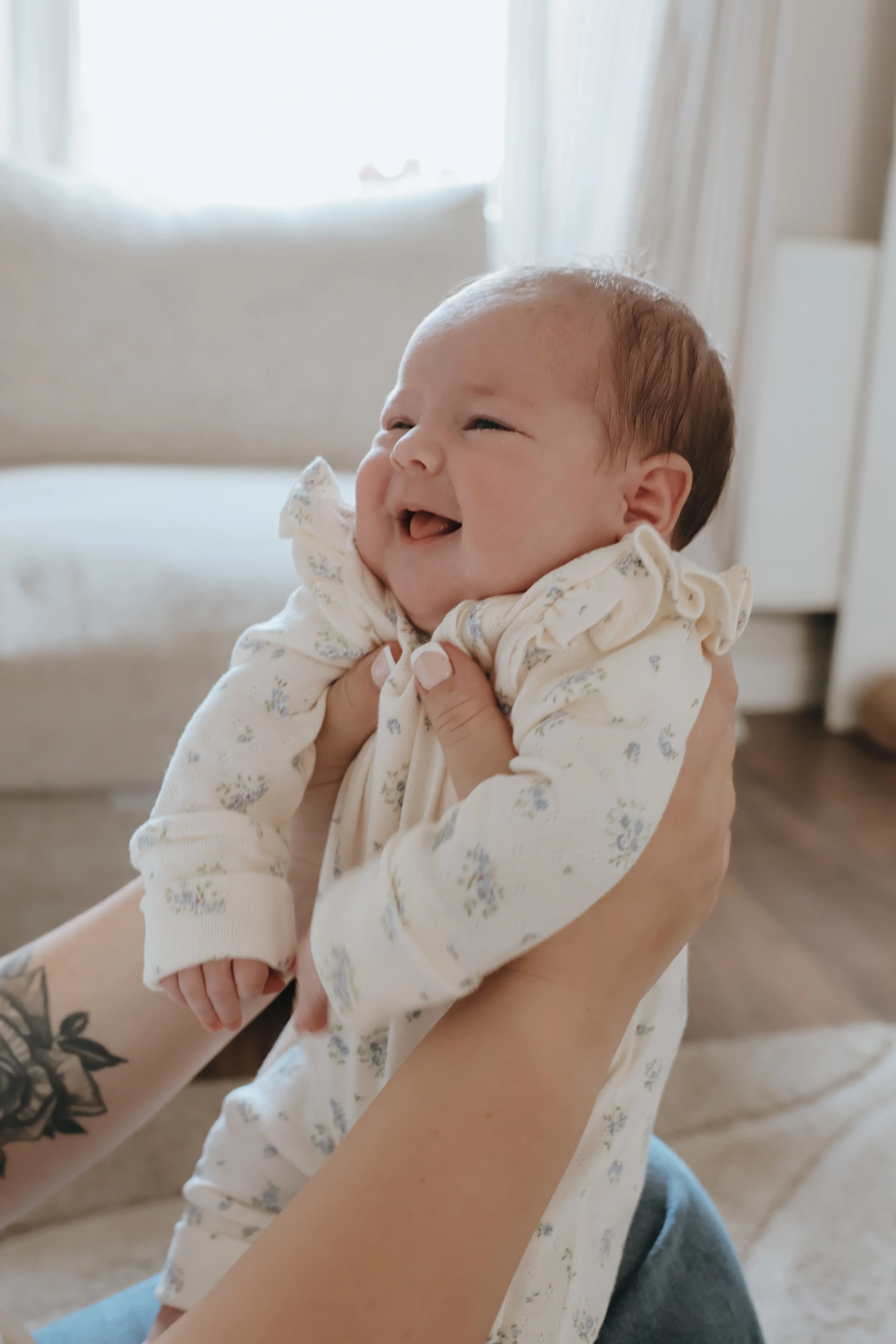 A baby with light skin, brown hair, and a big smile, wearing a light-colored floral outfit, being held by a person with tattoos on their arm.