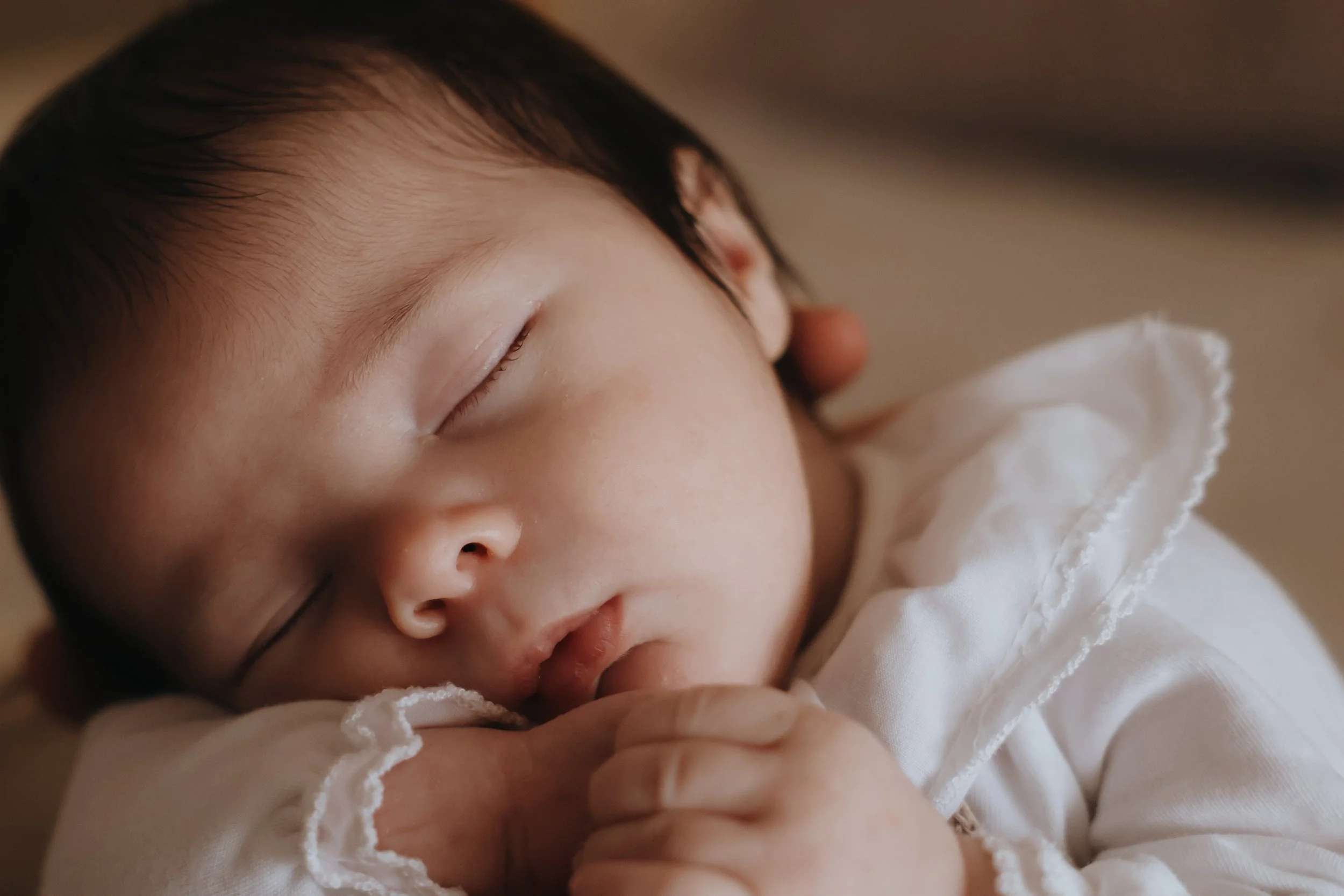 Close-up of a sleeping baby with dark hair, resting peacefully with hands near the face, wearing a white garment with ruffled edges.
Newborn photography, southampton