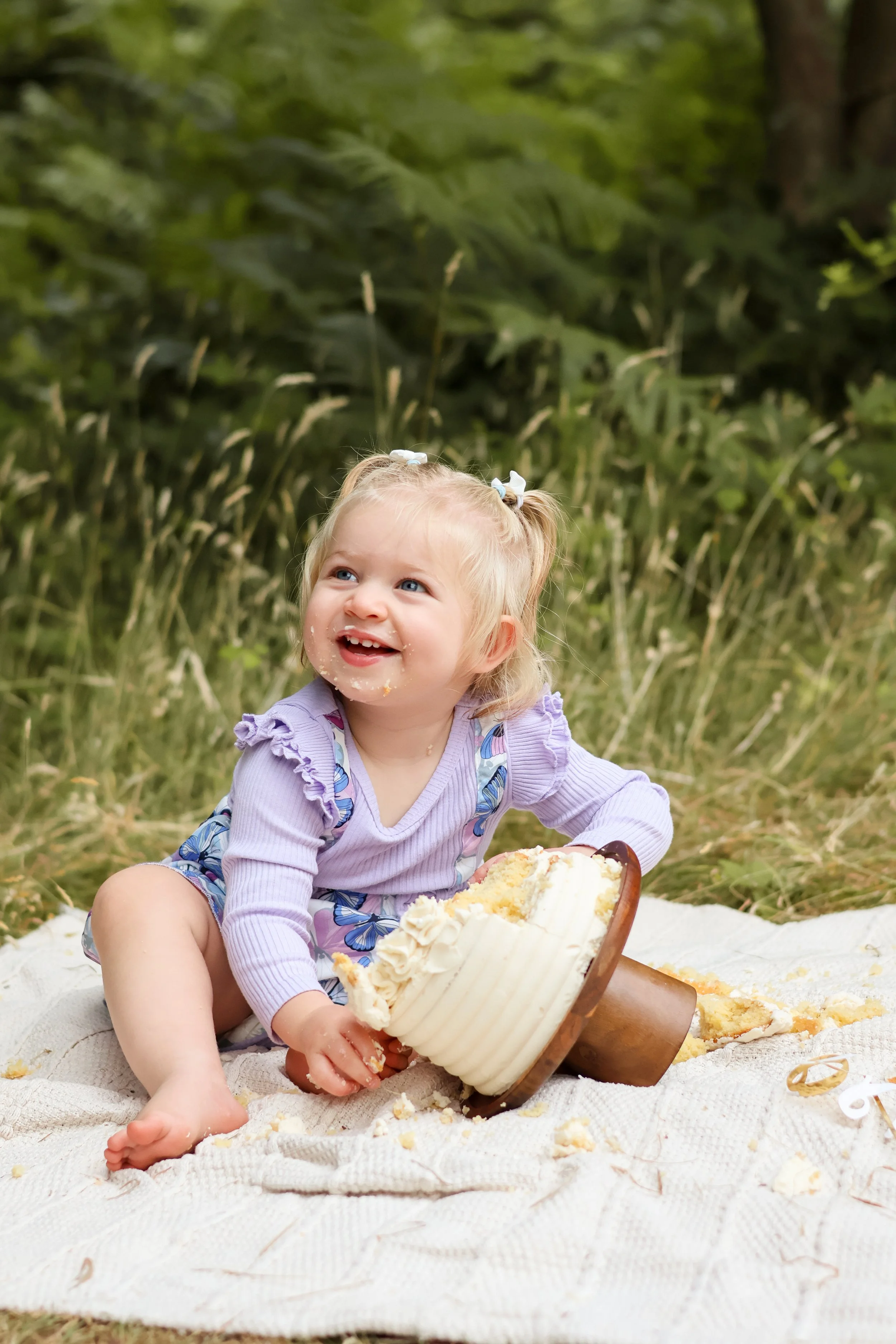 A young girl with blonde hair tied in pigtails, sitting on a blanket outdoors, smiling, with a partially eaten cake in front of her, surrounded by a green forest background. cake smash, first birthday photography, southampton 