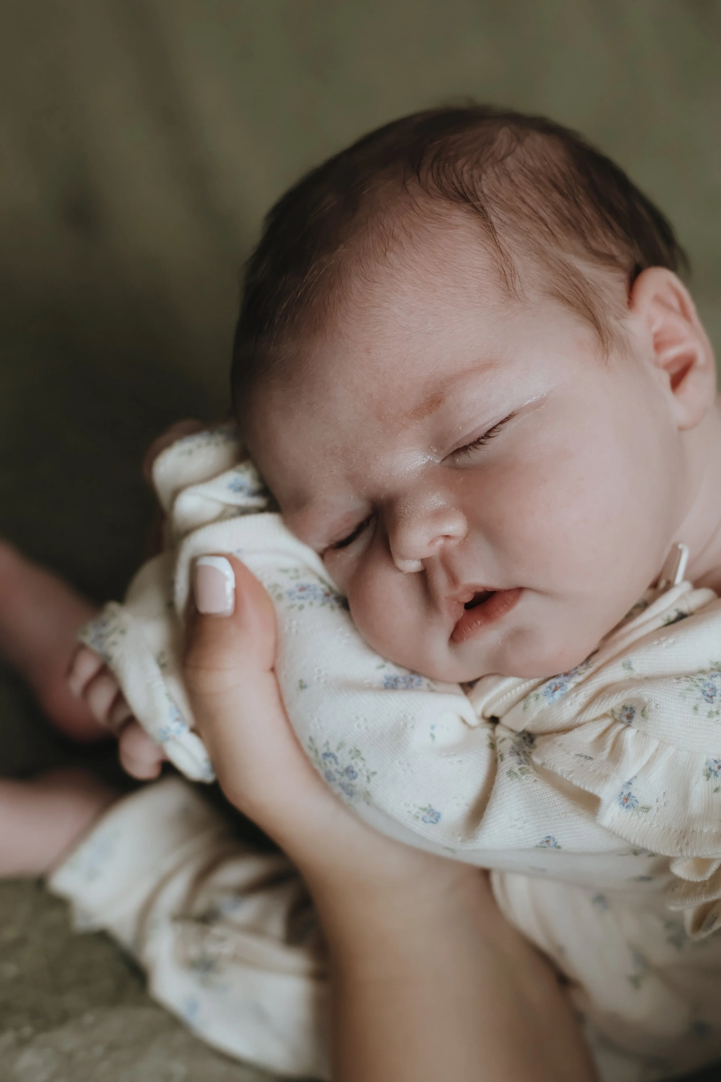 A close-up of a young child sleeping peacefully, held gently by someone, wearing a light-colored floral-patterned outfit, with a soft, blurred background.