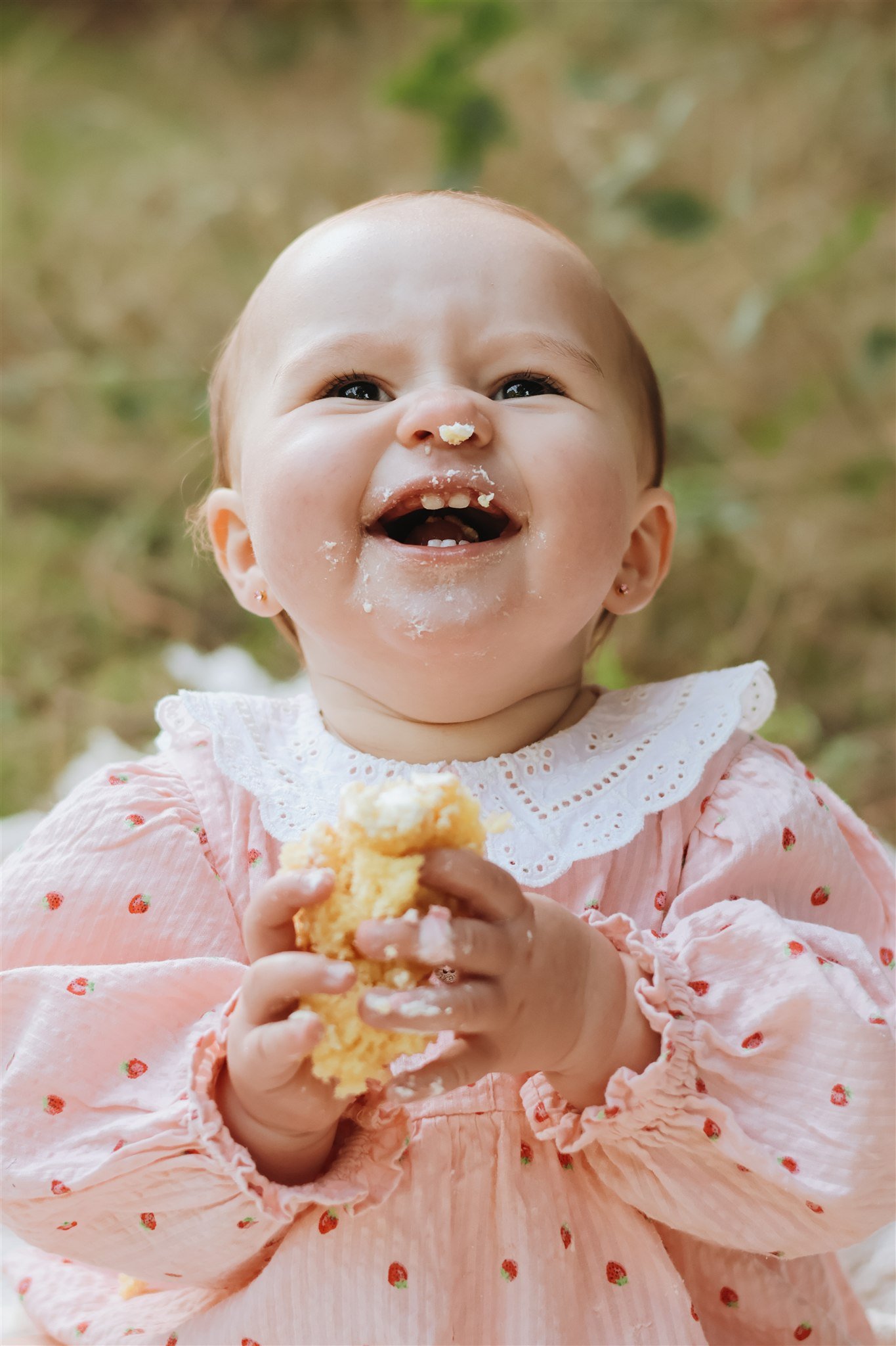 A smiling toddler girl with light skin and short, light-colored hair, wearing a pink dress with a white lace collar and small strawberry prints, holding a piece of cake in her hands, with cake crumbs on her face. cake-smash session, southampton