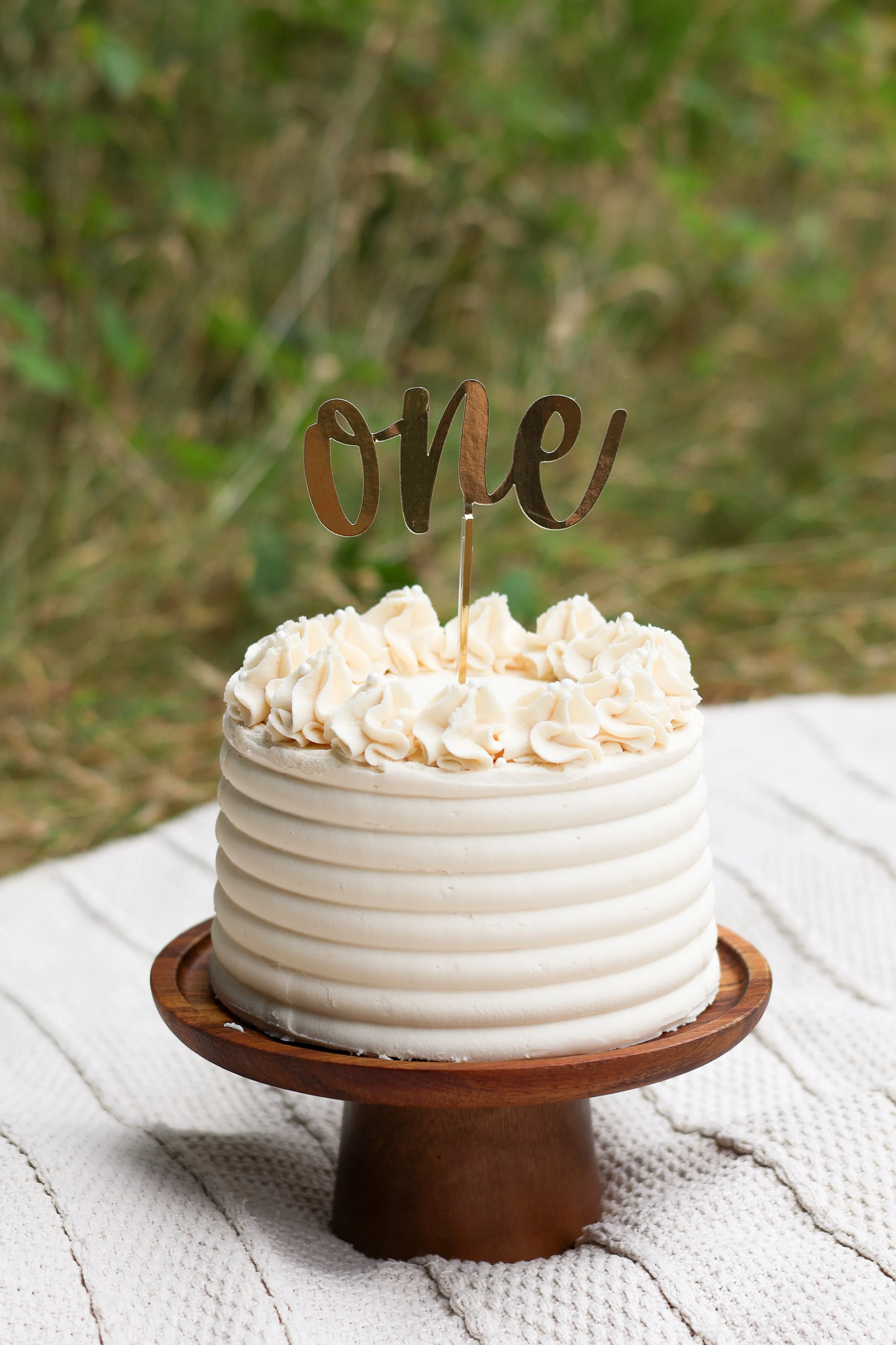 A white layer cake with ridged sides and white frosting rosettes, topped with a metallic cake topper that says 'one', sitting on a wooden cake stand on a white textured cloth outdoors.