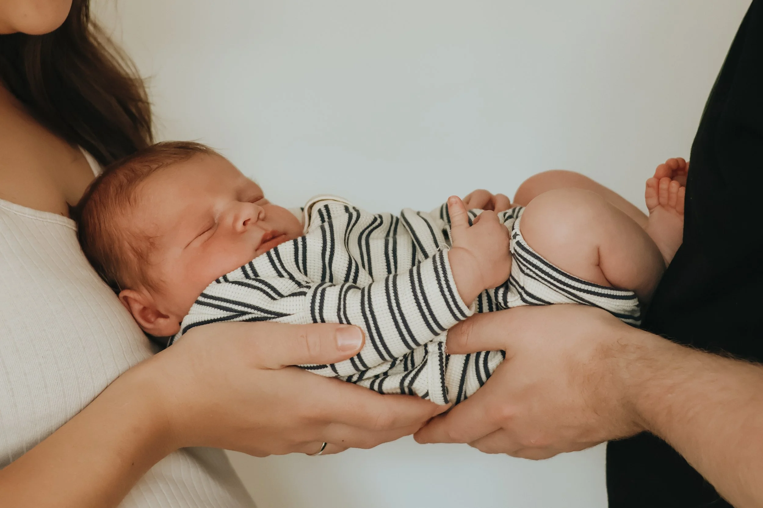 A newborn baby sleeping peacefully on an adult's chest, being gently held by two adults with one hand supporting the baby's back and the other supporting the baby's legs.