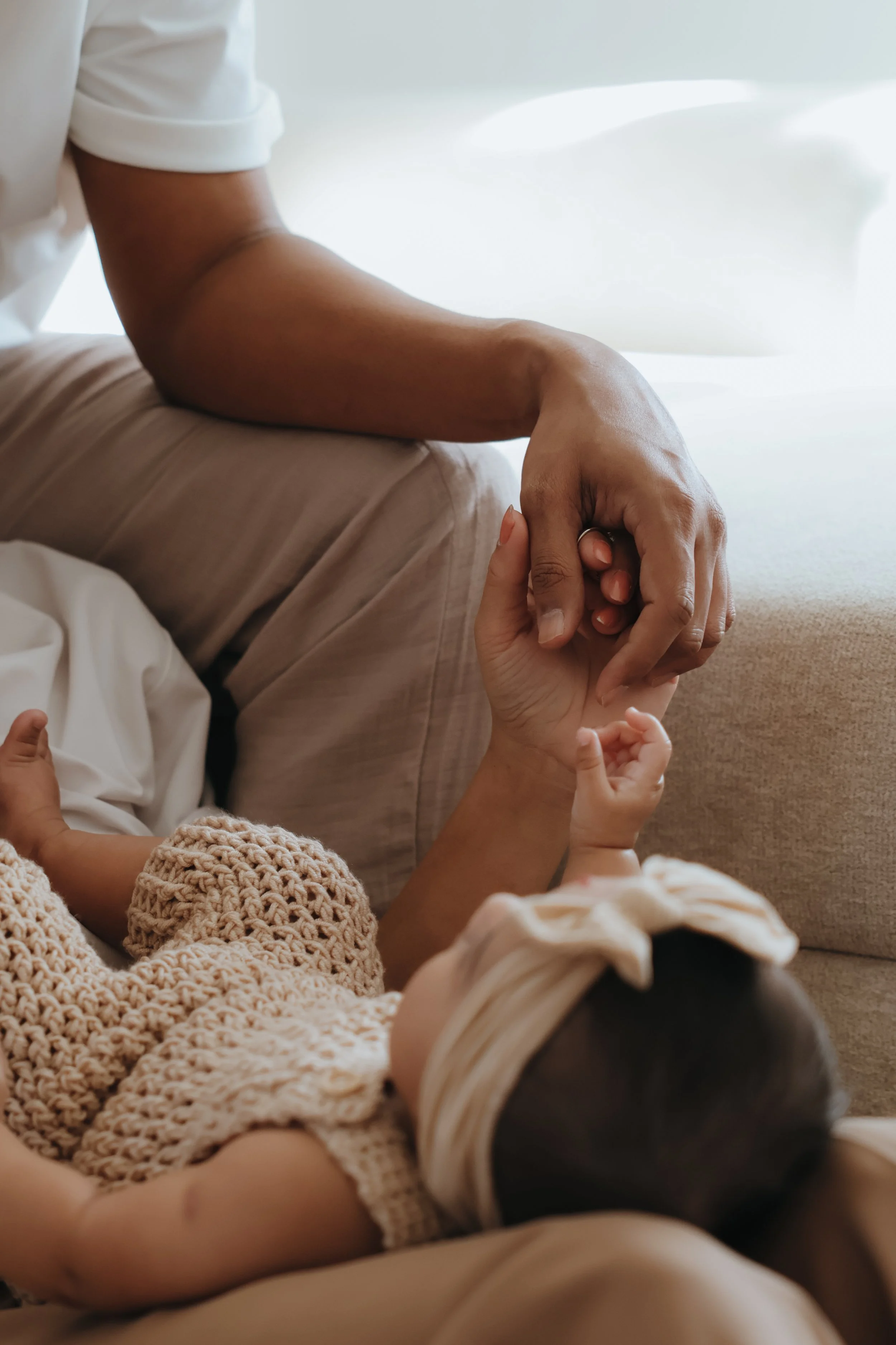 A person holding a baby's hand while sitting on a beige couch.