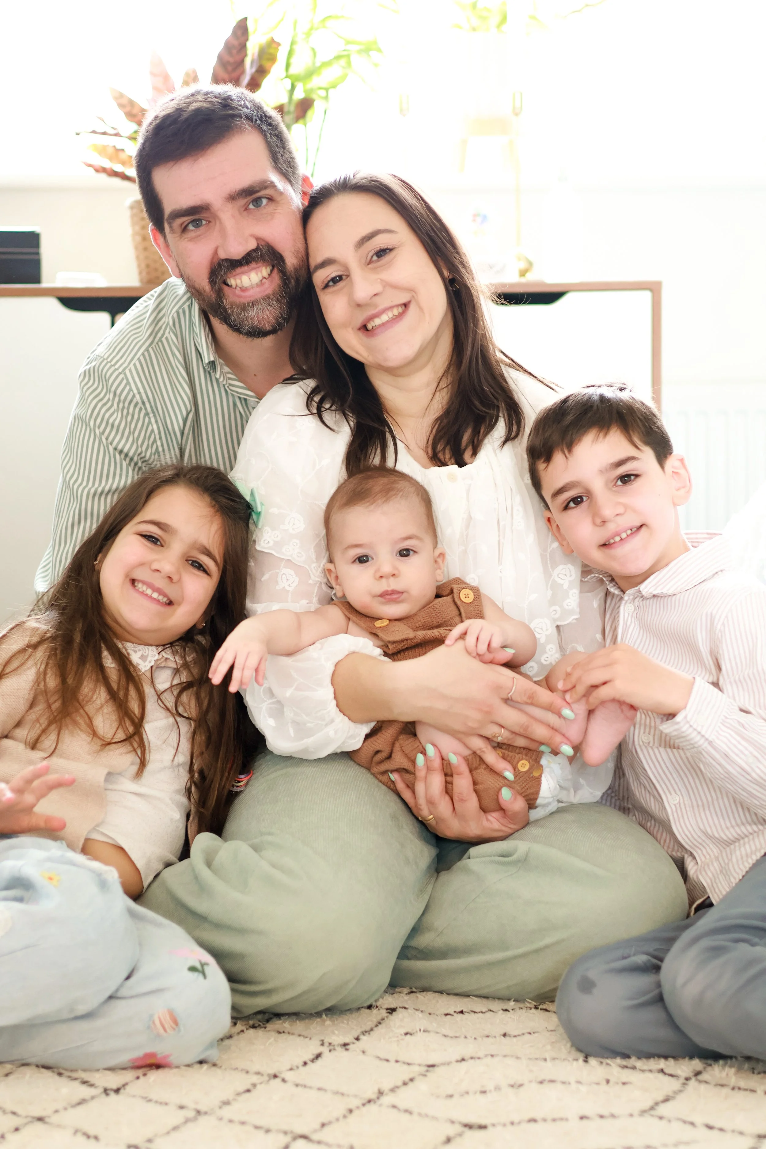 A family of six sitting on a carpet, smiling and hugging inside a bright room with plants in the background.