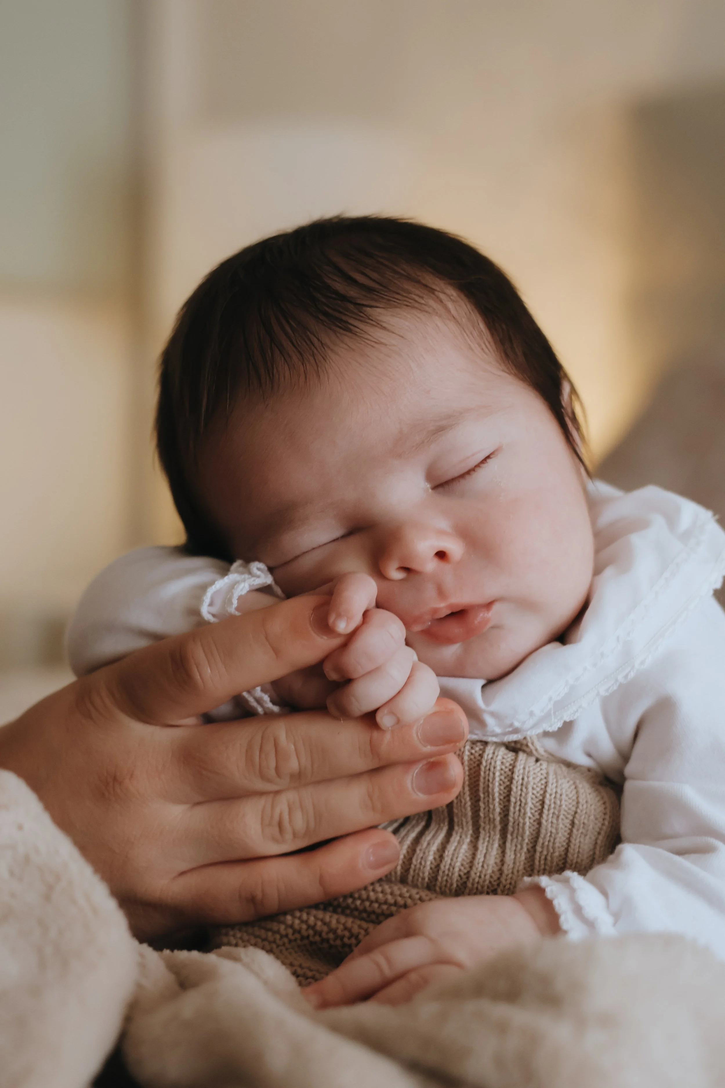 A sleeping baby with dark hair, being gently held by an adult's hand, resting on a soft blanket.