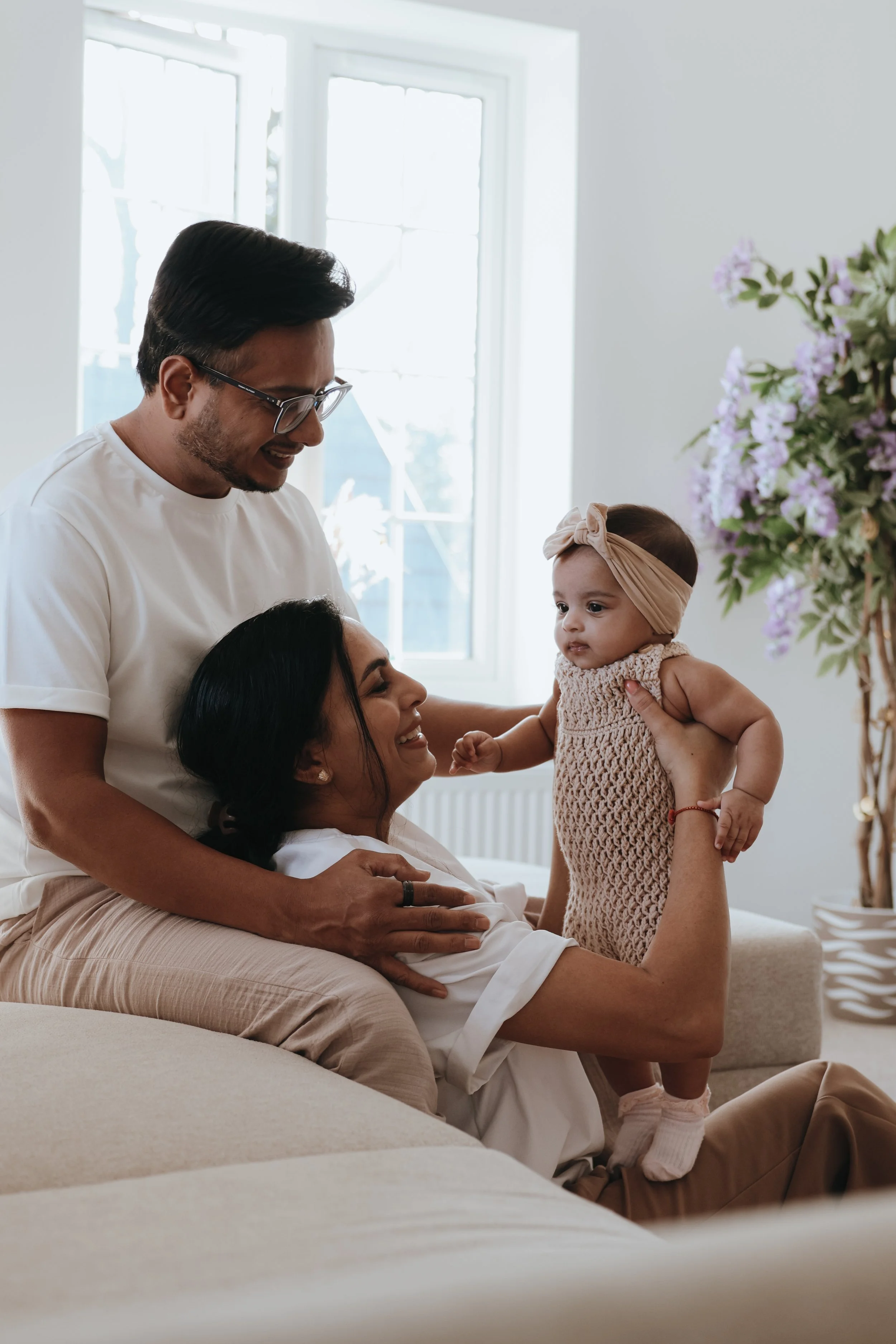 A happy family of three enjoying time together in a bright living room with large windows and a flower arrangement, featuring an adult woman sitting on a couch holding a baby girl, while a man stands behind them, all smiling and engaging with each other.