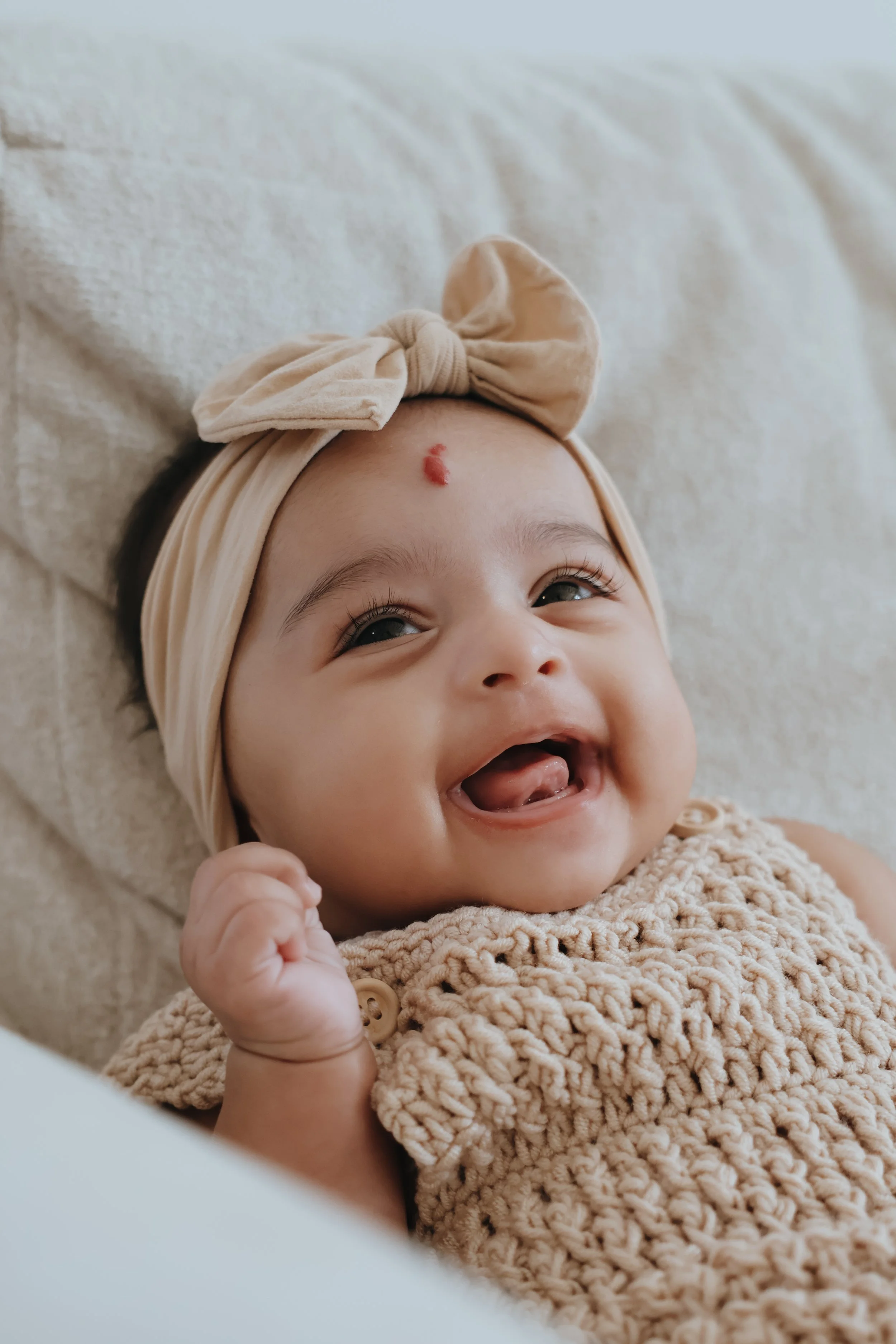 Smiling baby girl lying on a soft surface, wearing a beige knitted outfit and a headband with a bow, with a small red bump on her forehead.