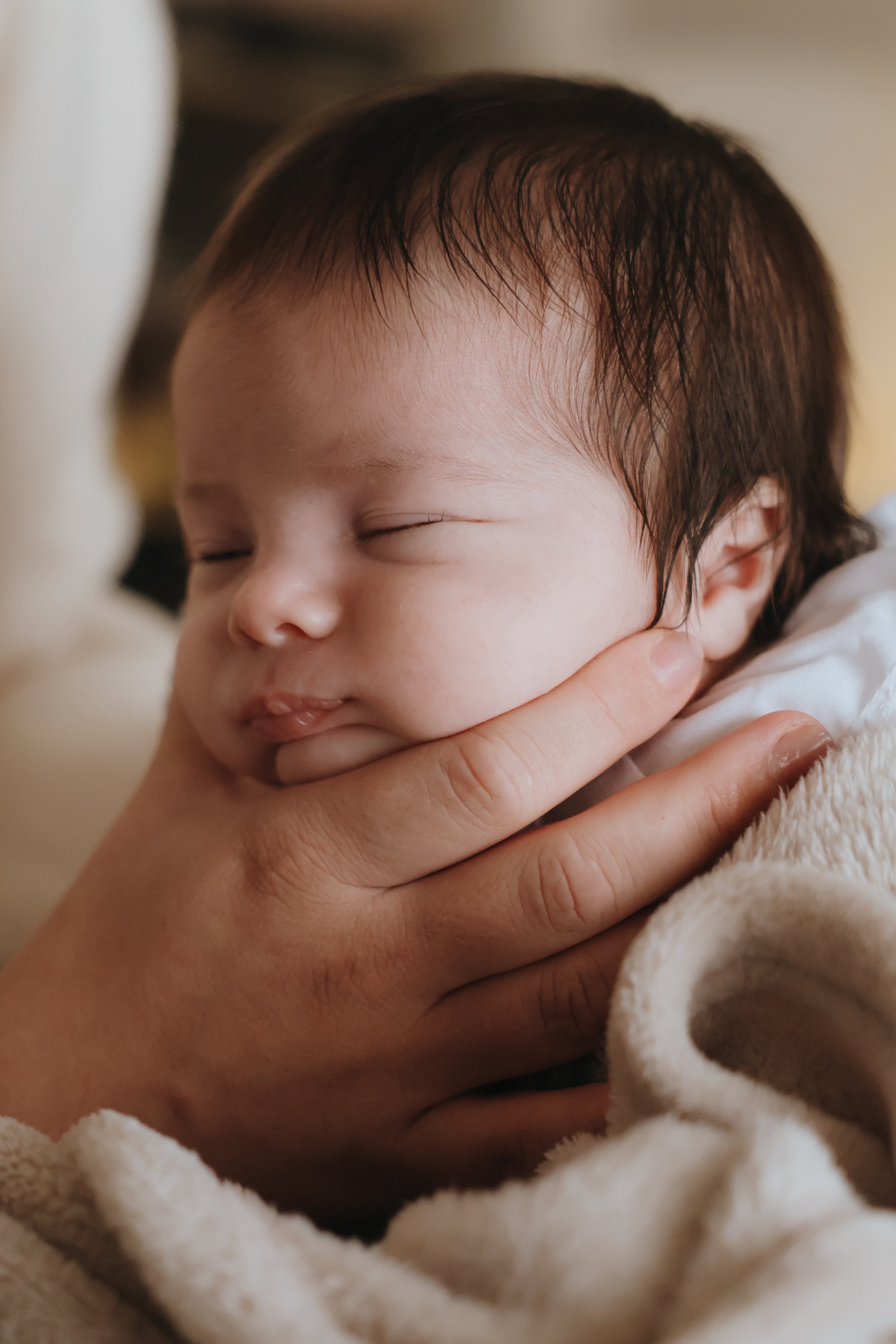 A close-up of a sleeping baby with dark hair, being gently held by an adult's hand under its chin. Newborn photography southampton 