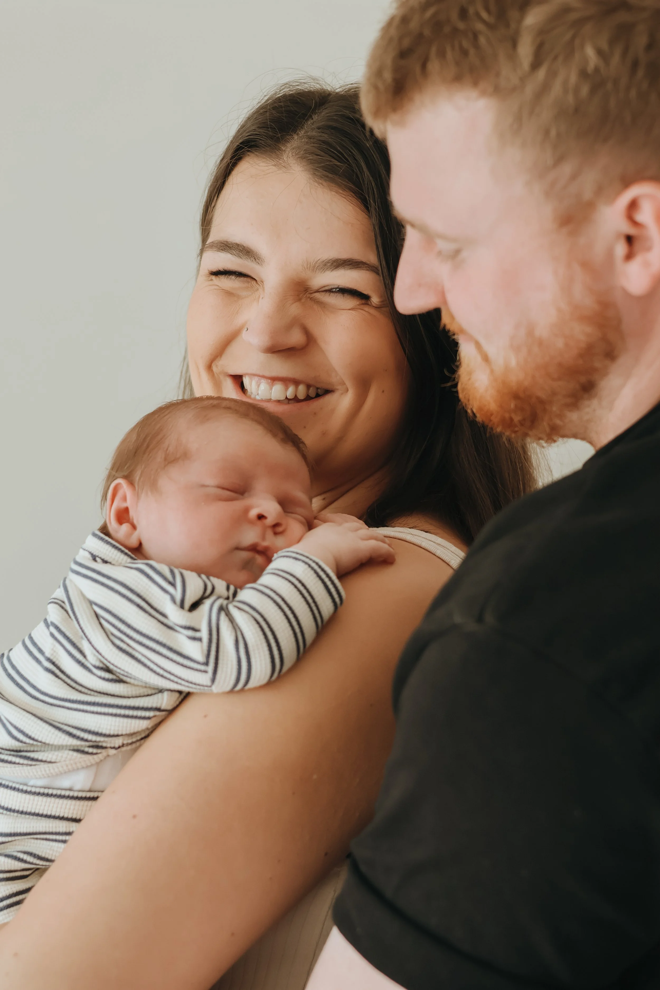 A smiling woman holding a sleeping baby on her shoulder, with a man leaning in close, both showing affection and warmth.