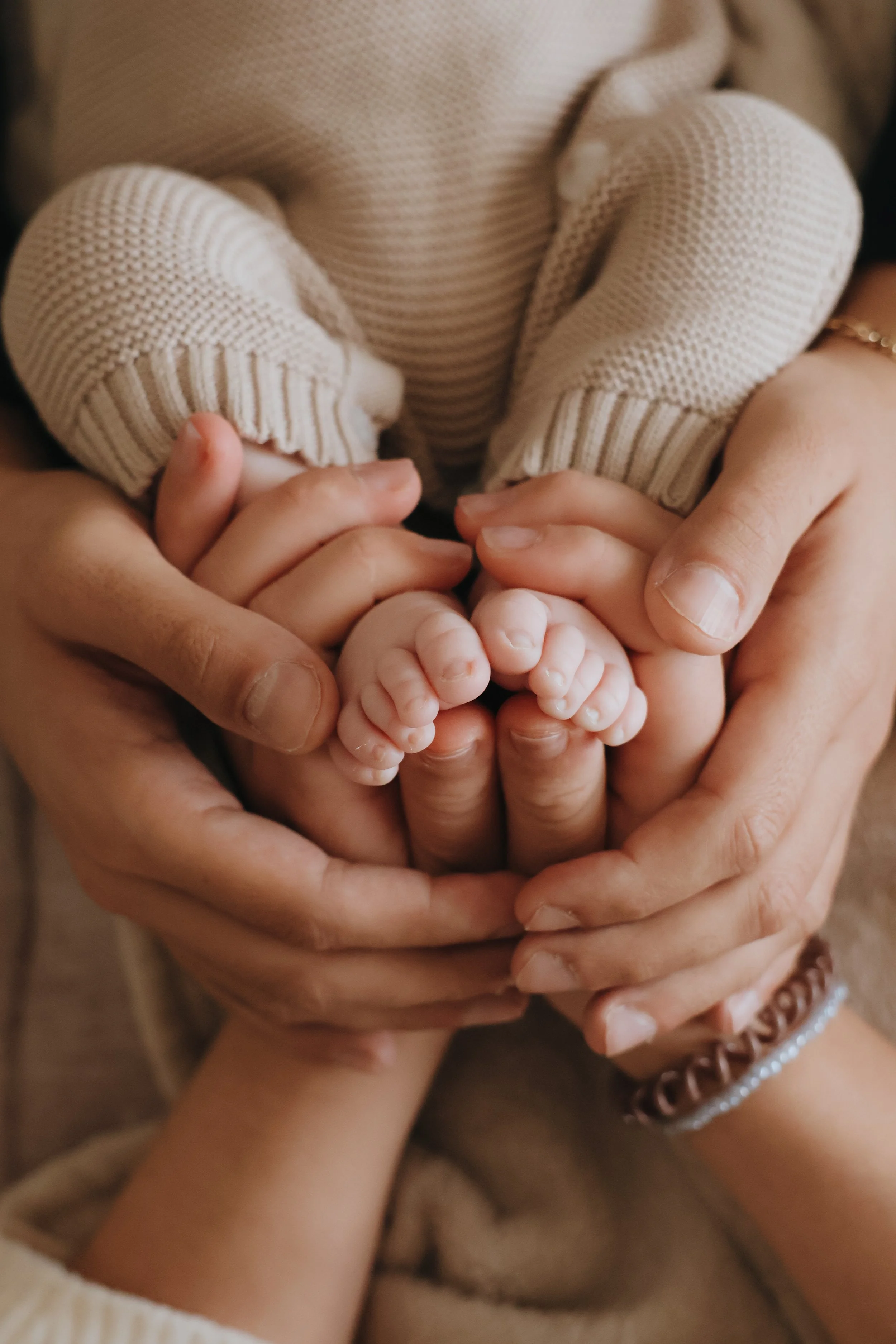 Multiple hands, including an adult's and a child's, gently cradling tiny baby feet, with the baby wearing cream-colored socks. In southampton
