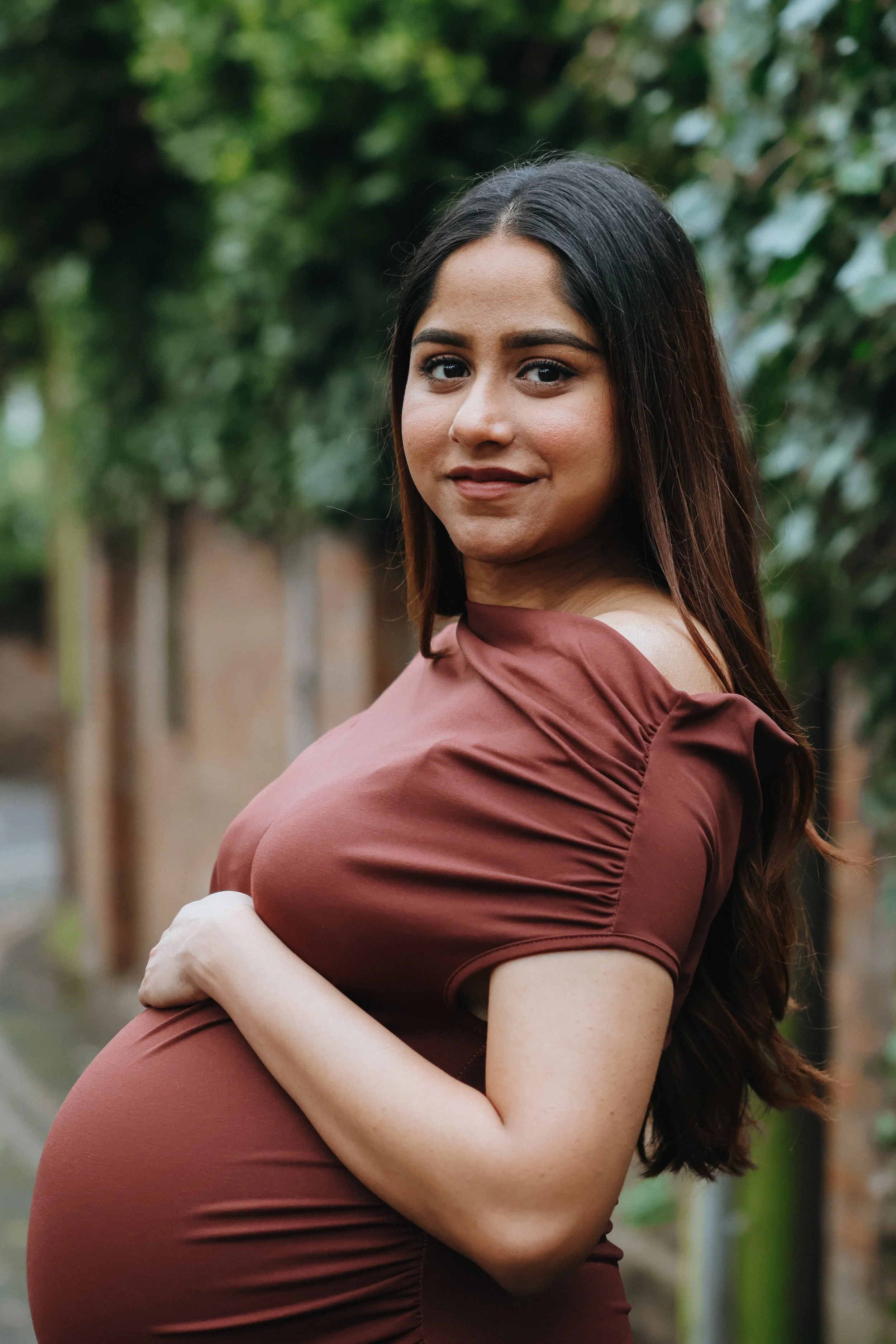 A pregnant woman with long dark hair and medium skin tone, wearing a brown dress, standing outdoors in front of green trees, looking at the camera with a slight smile.