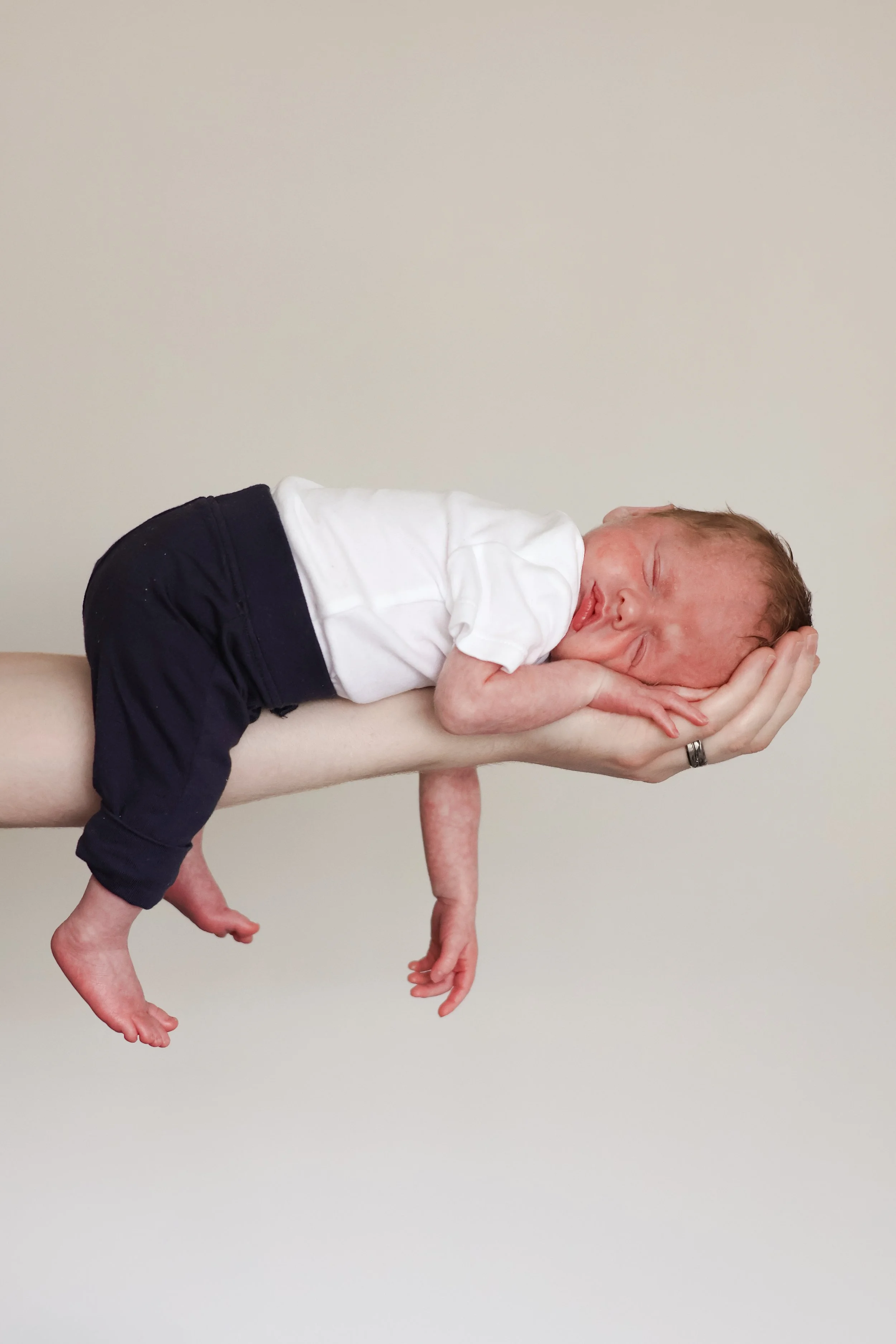 A newborn baby with reddish hair is lying on an adult's arm, wearing a white shirt and dark pants, against a plain background.
