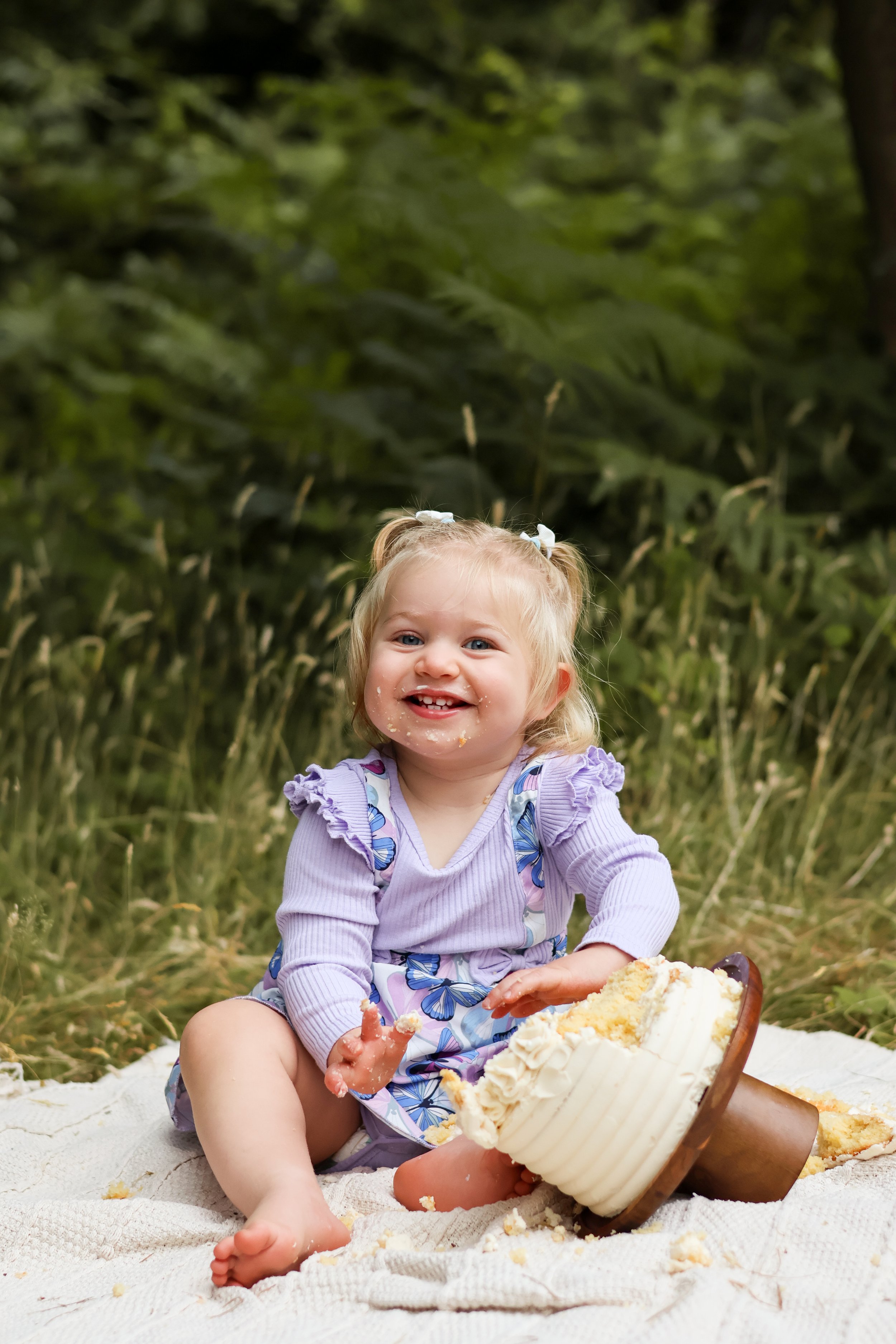 A young girl with blonde hair in pigtails, wearing a purple dress with butterfly patterns, sitting on a white blanket outdoors. She is happily playing with a cake that has toppled over, with cake crumbs and frosting on her face and hands. The backgro