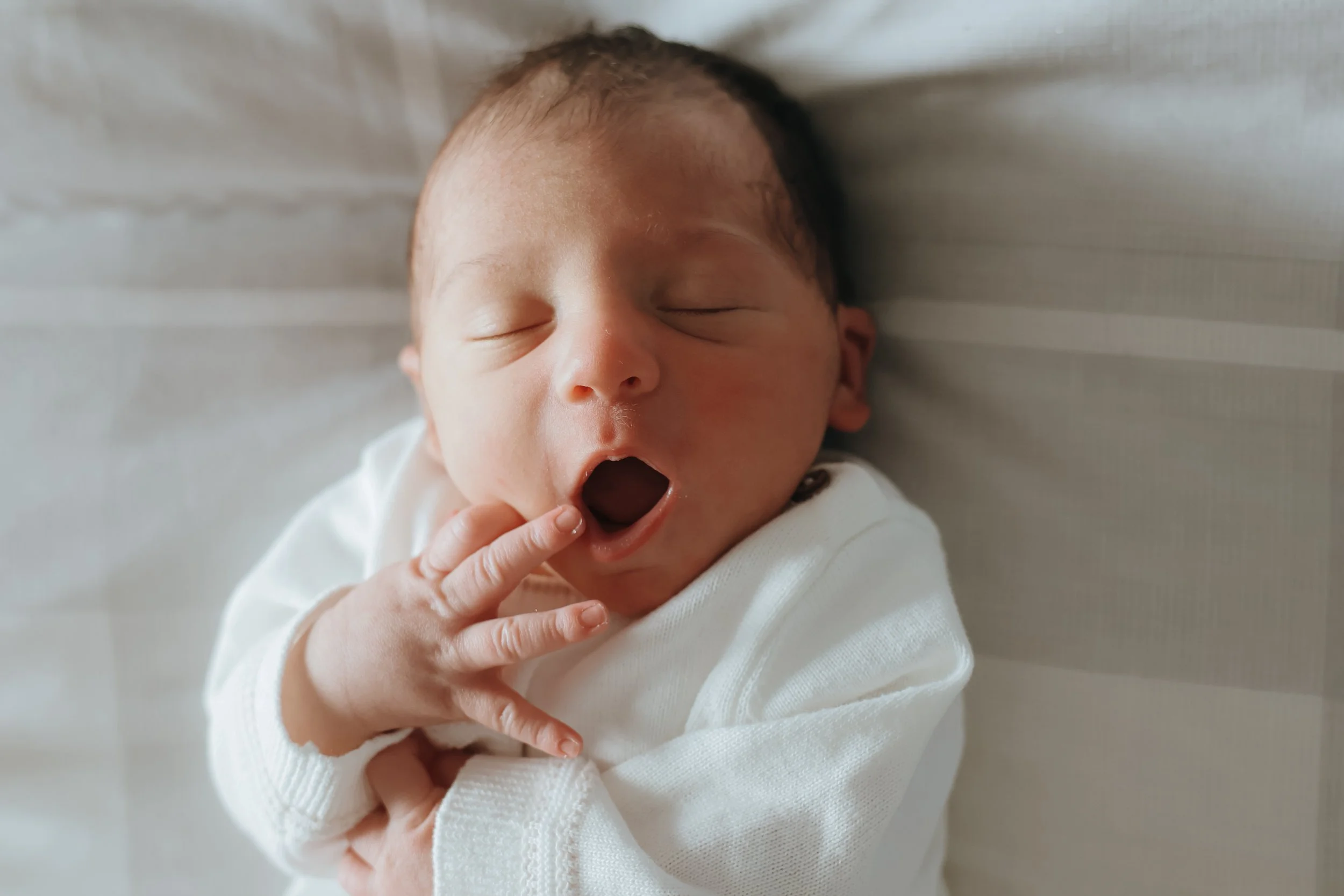 A newborn baby yawning with fingers near the mouth, lying on a white surface. Newborn photography, bournemouth 