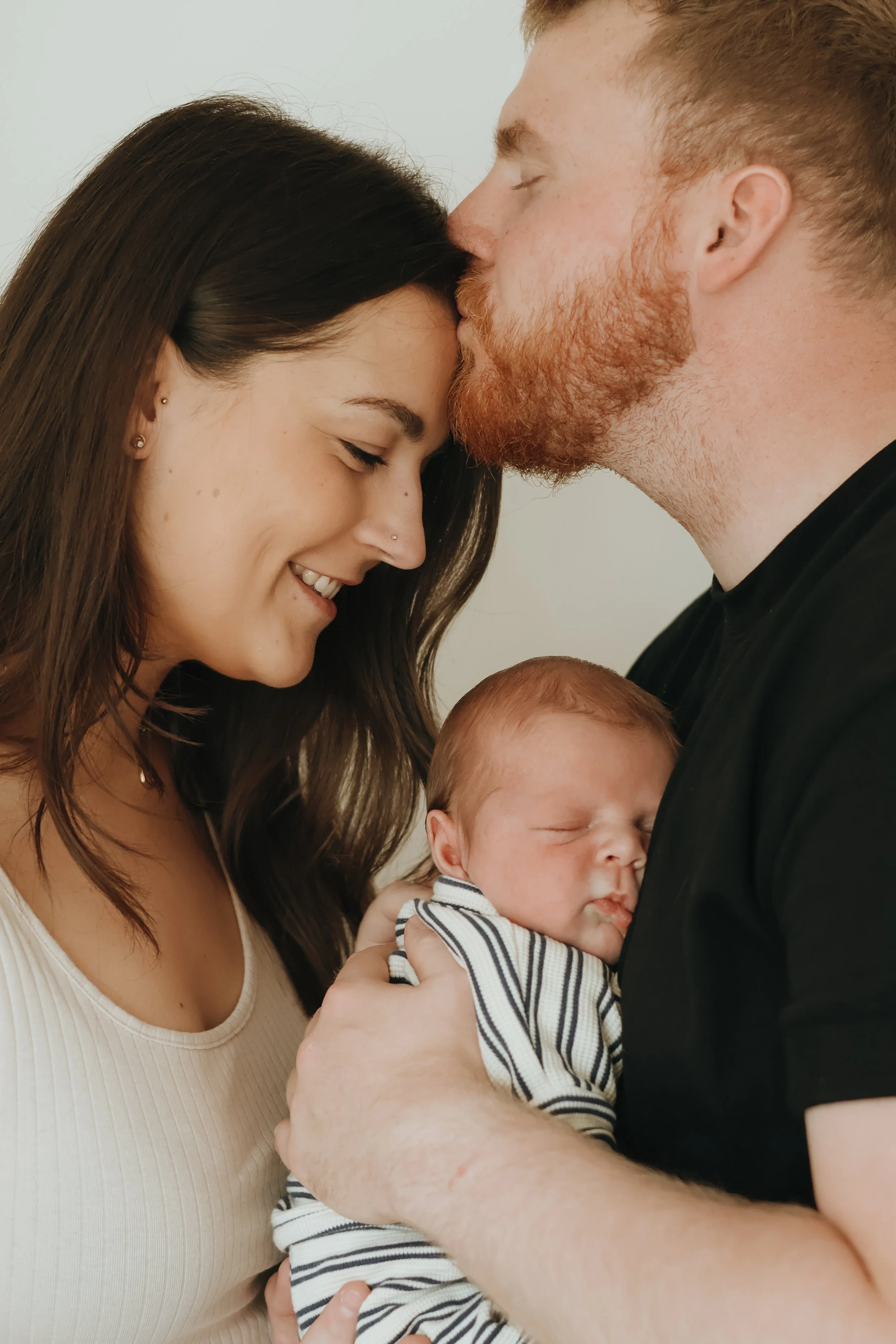A family with a mother, father, and their newborn baby. The father is kissing the mother's forehead while she smiles with her eyes closed, and the baby is peacefully sleeping in the father's arms.