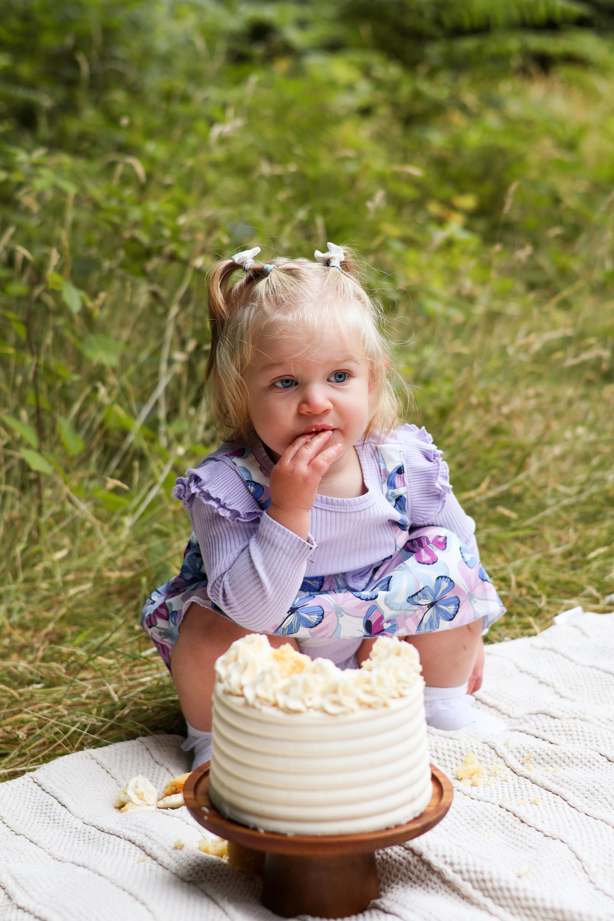 A young girl with blonde hair in pigtails and blue eyes squats on a picnic blanket outdoors, touching her lips, with a partially eaten birthday cake in front of her, surrounded by green foliage.