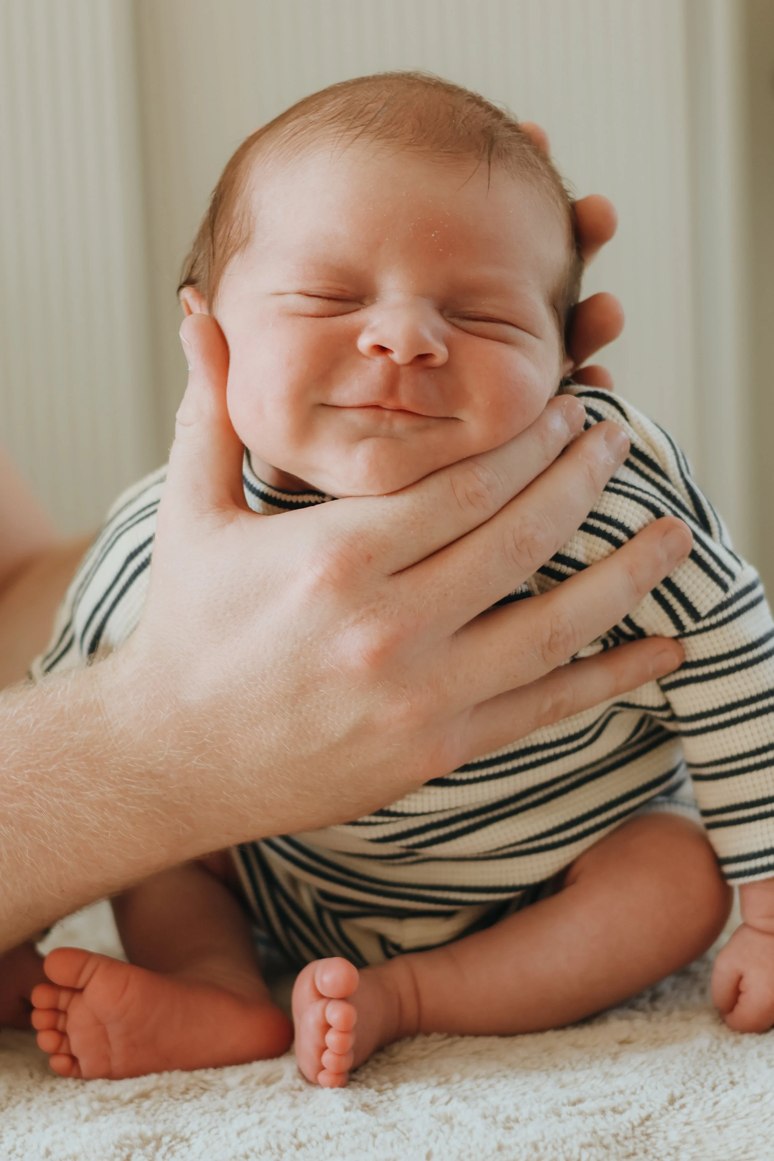 A happy newborn baby with closed eyes, being gently held by an adult hand, on a soft surface.