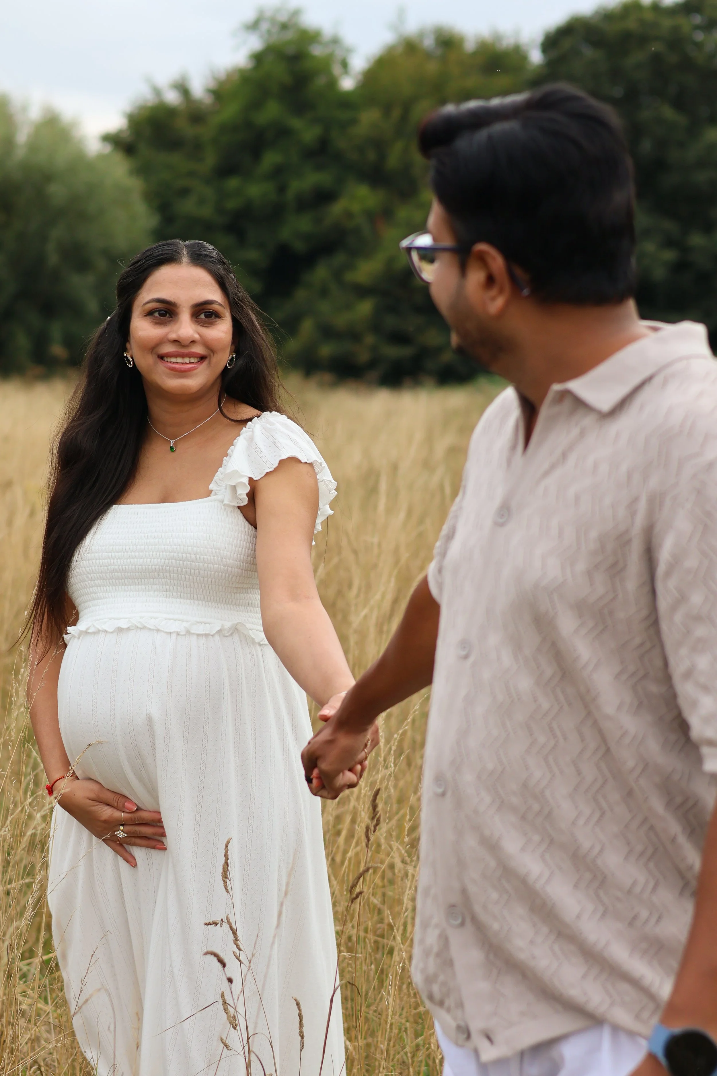 A pregnant woman in a white dress holding her belly and smiling while holding hands with a man in a short-sleeved shirt in a field with tall grass and trees in the background.