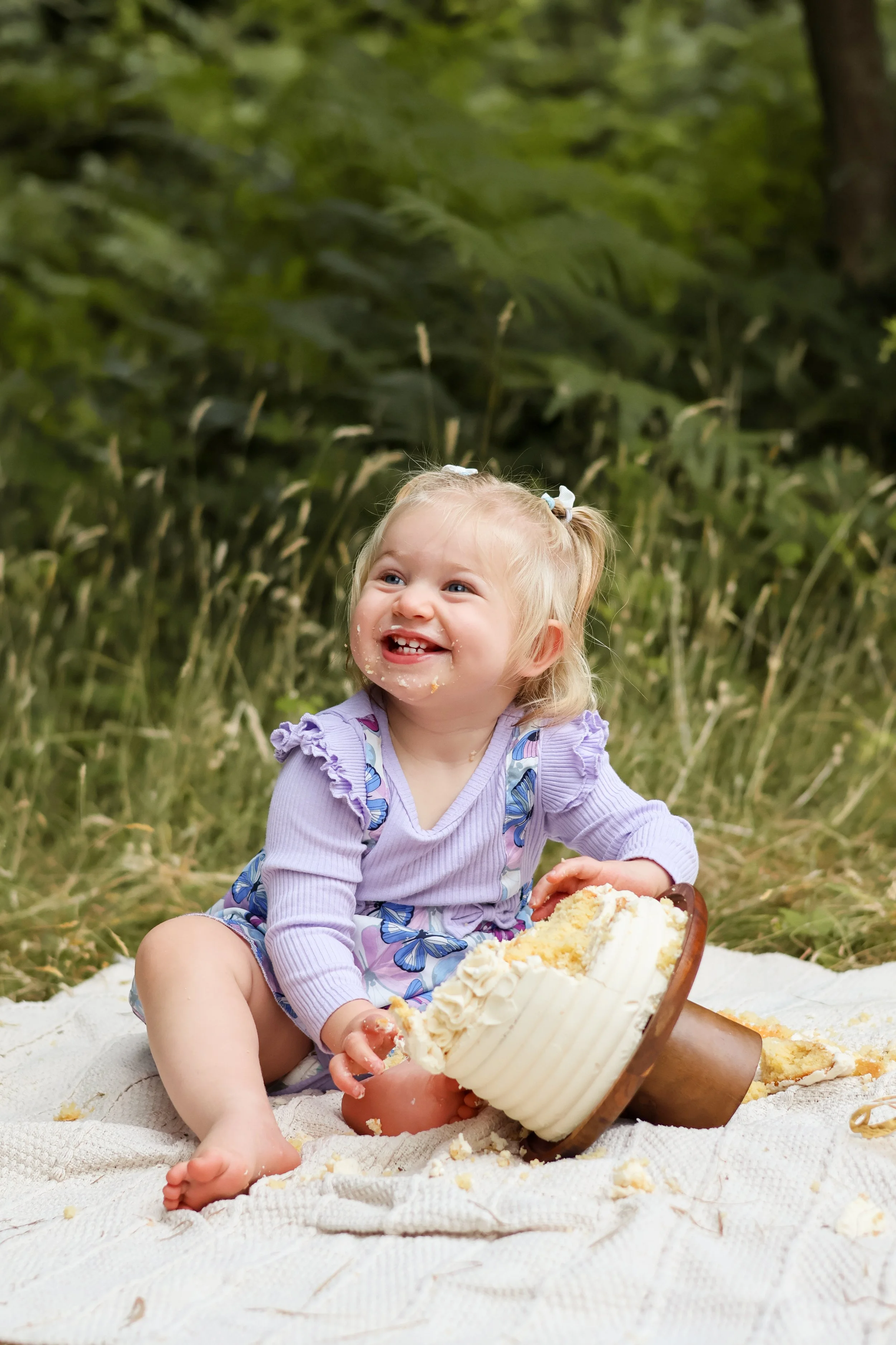 A young girl with blonde hair tied in pigtails, wearing a purple dress with butterfly patterns, sitting on a white blanket outdoors in a grassy area, eating cake with white frosting, smiling and looking happy.