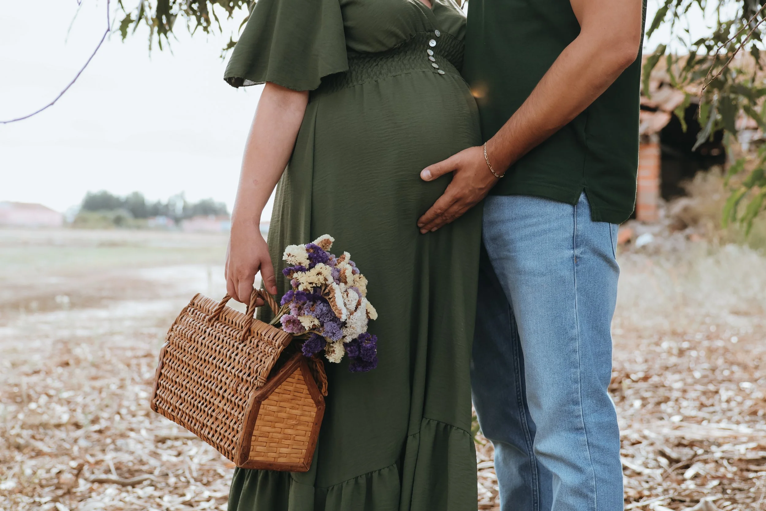 A pregnant woman in an olive green dress holding a basket with flowers while a man in a green shirt and jeans touches her belly with his hand outdoors. maternity photography outdoor southampton