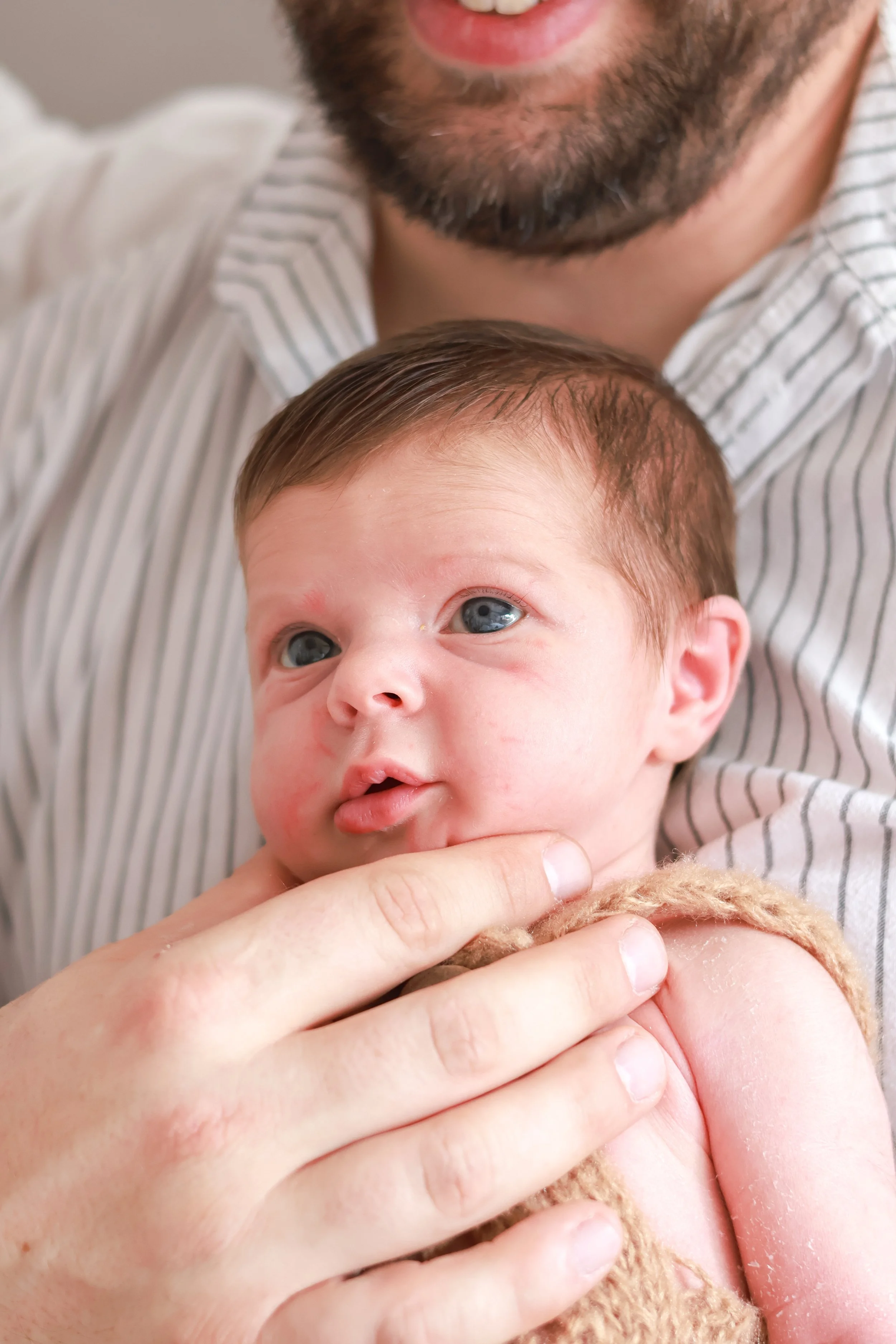 Close-up of a father holding a newborn baby with blue eyes, visible lips, and light skin, while gently touching the baby's cheek with his hand.