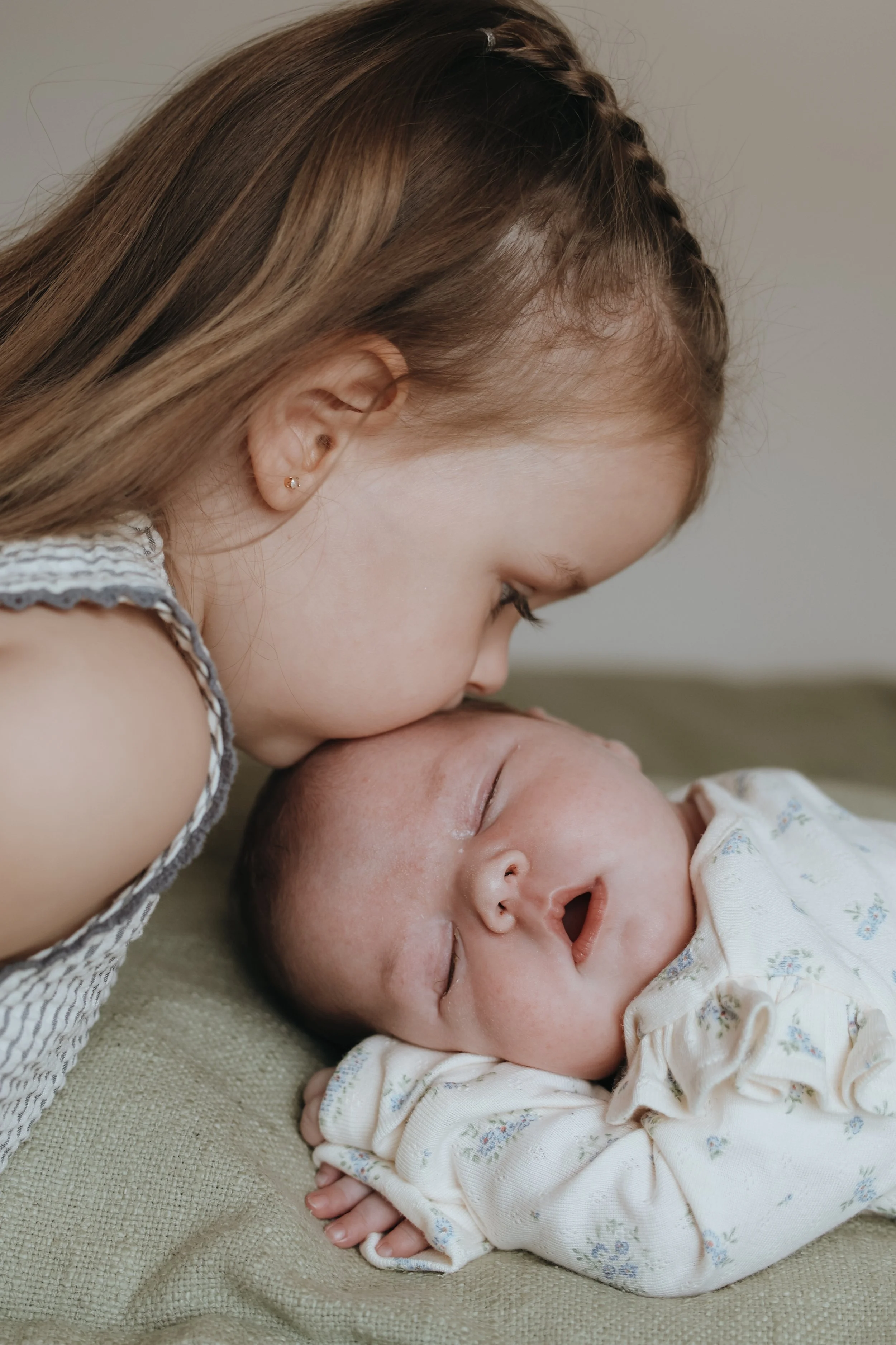 A young girl with light brown hair and a small earring gently kisses a newborn baby who is lying on a soft surface. The baby is asleep, wearing a cream-colored onesie with small floral patterns, and has a peaceful expression.